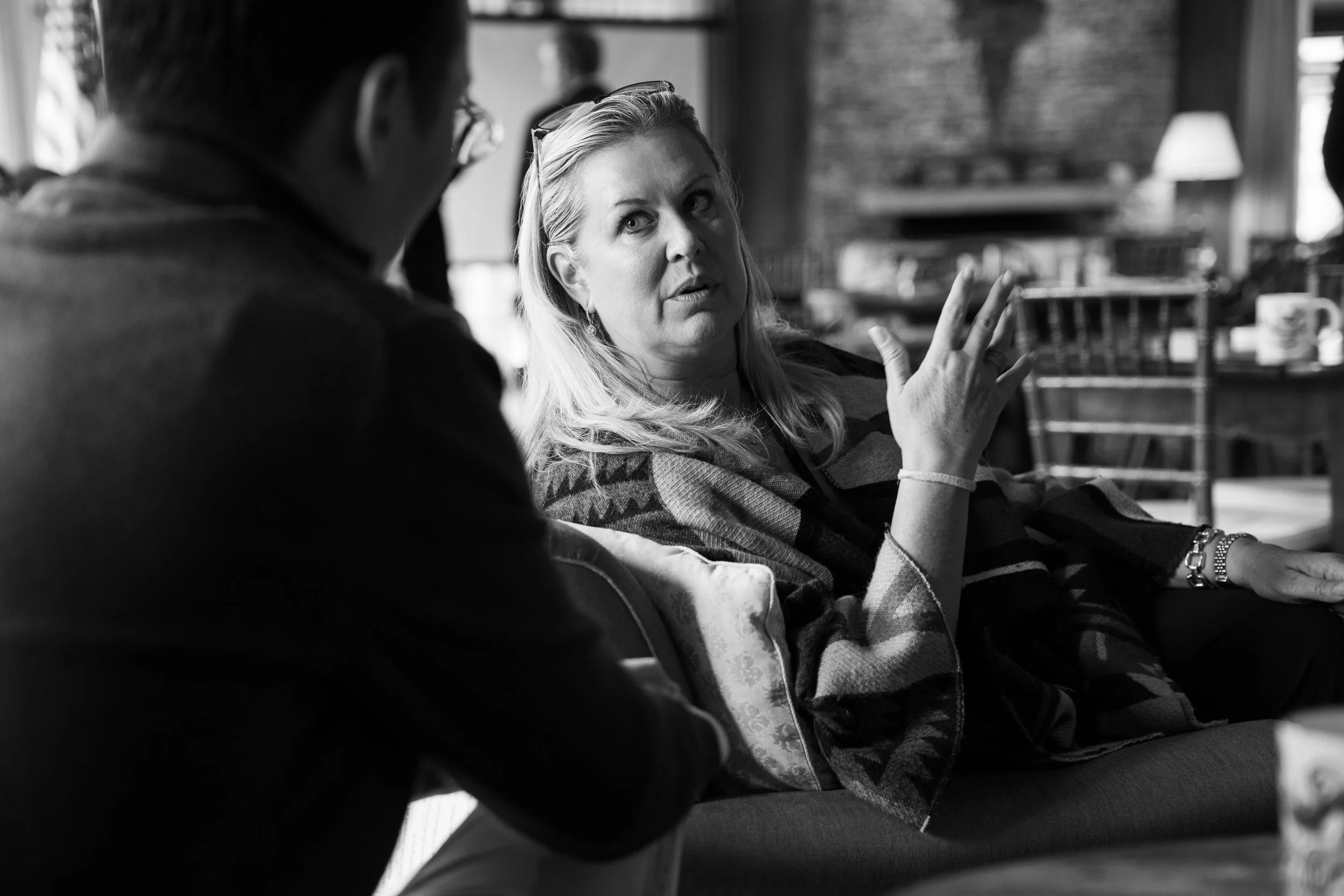 A woman with blonde hair and a striped sweater talk to a man wearing glasses in a cozy indoor setting, with a wooden fireplace and ambient lighting in the background.