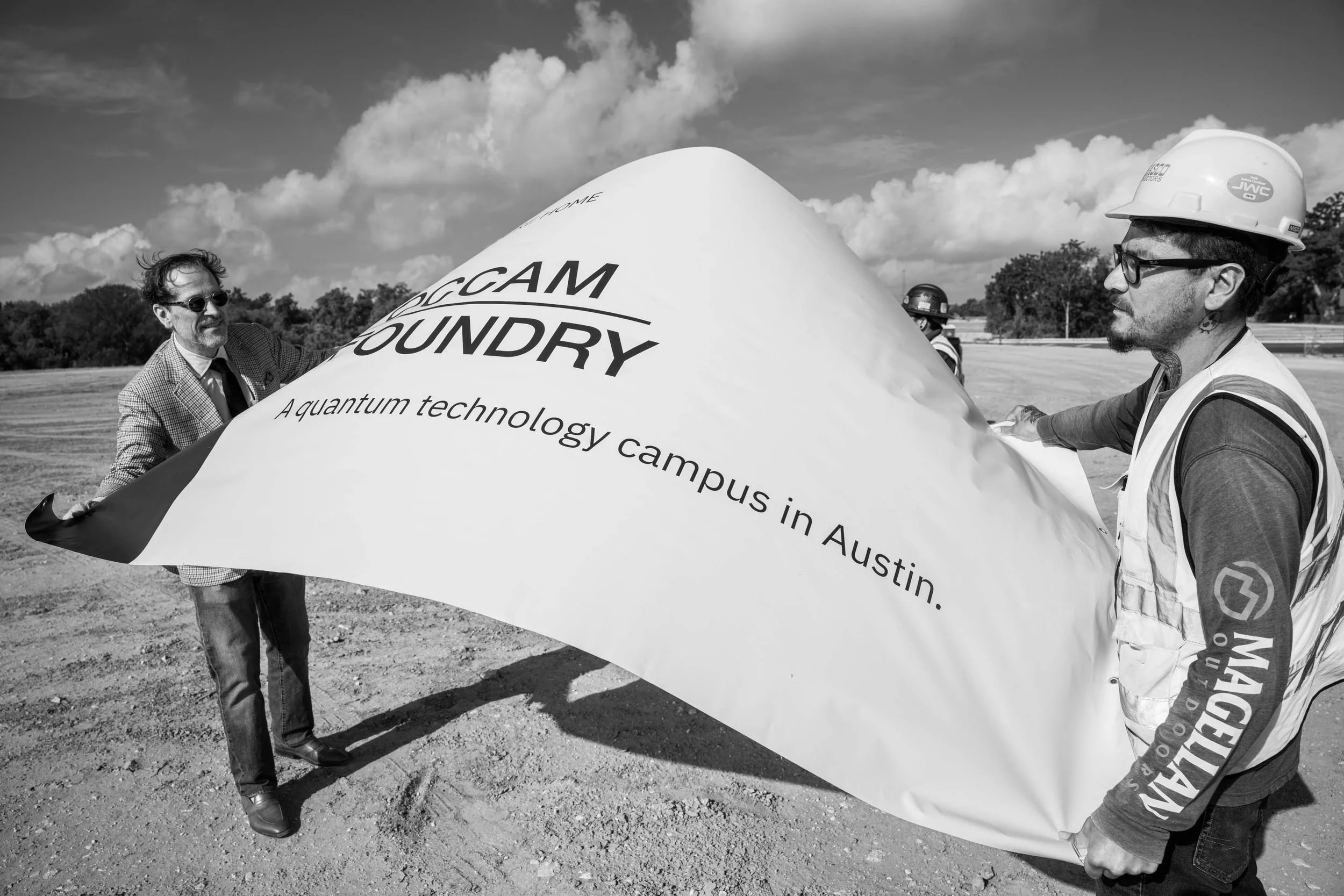 Two men holding a large banner that reads "OCCAM FOUNDRY" and "A quantum technology campus in Austin." outdoors on a sunny day with clouds in the sky.