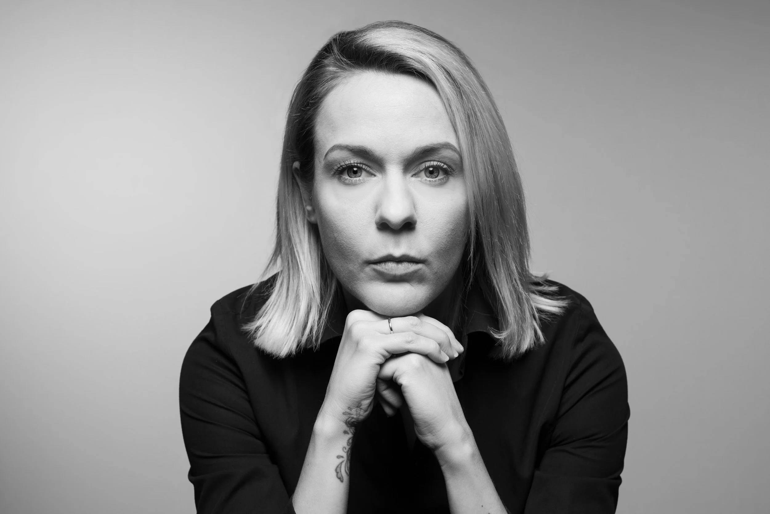 Black and white portrait of a woman with shoulder-length hair, wearing a dark top, holding her chin with both hands, looking directly at the camera against a plain background.