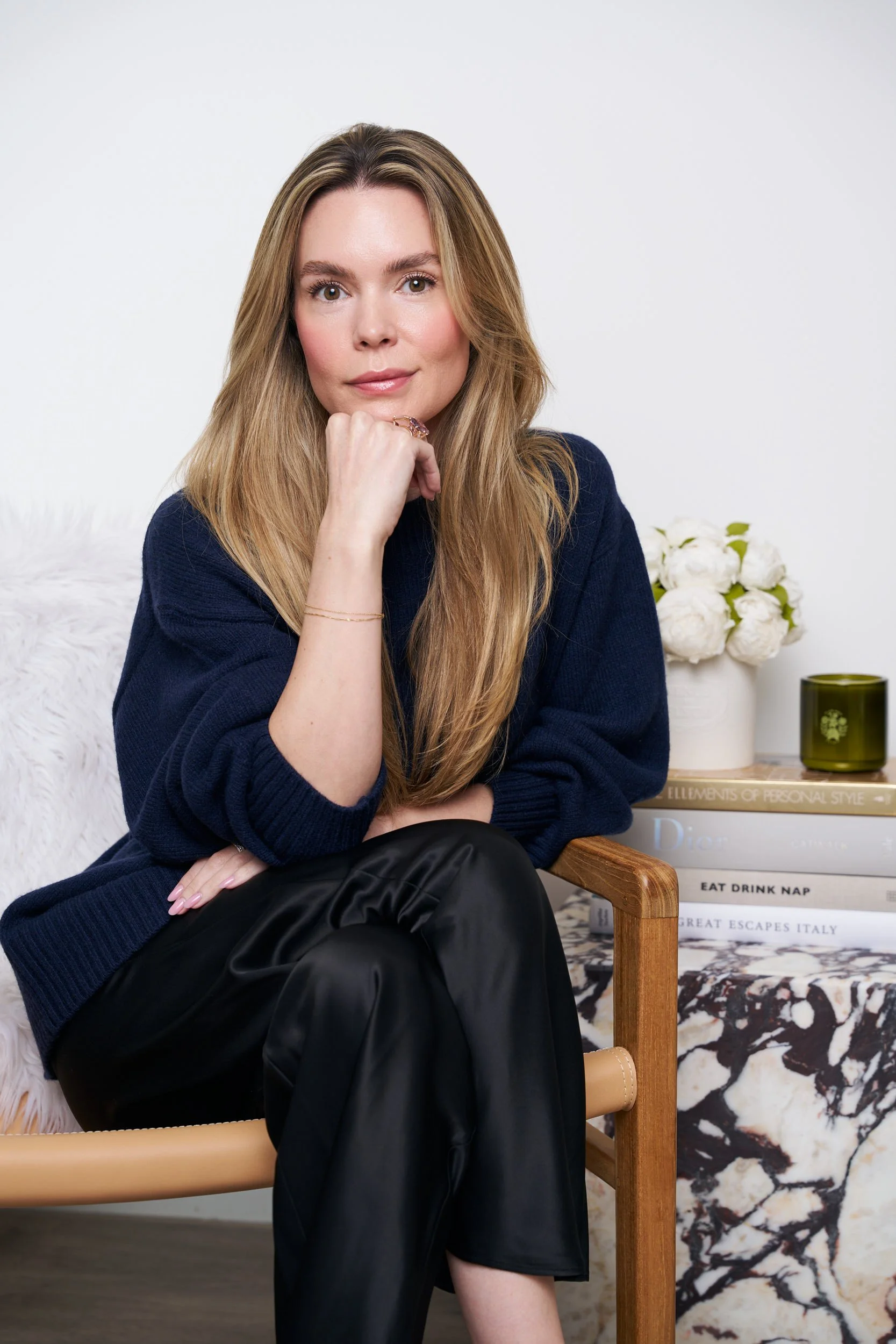 A woman with long, wavy blonde hair and fair skin is sitting on a chair with her right hand resting on her chin. She is wearing a navy blue sweater and black leather pants. In the background, there are white flowers in a vase, and stacked books on a 