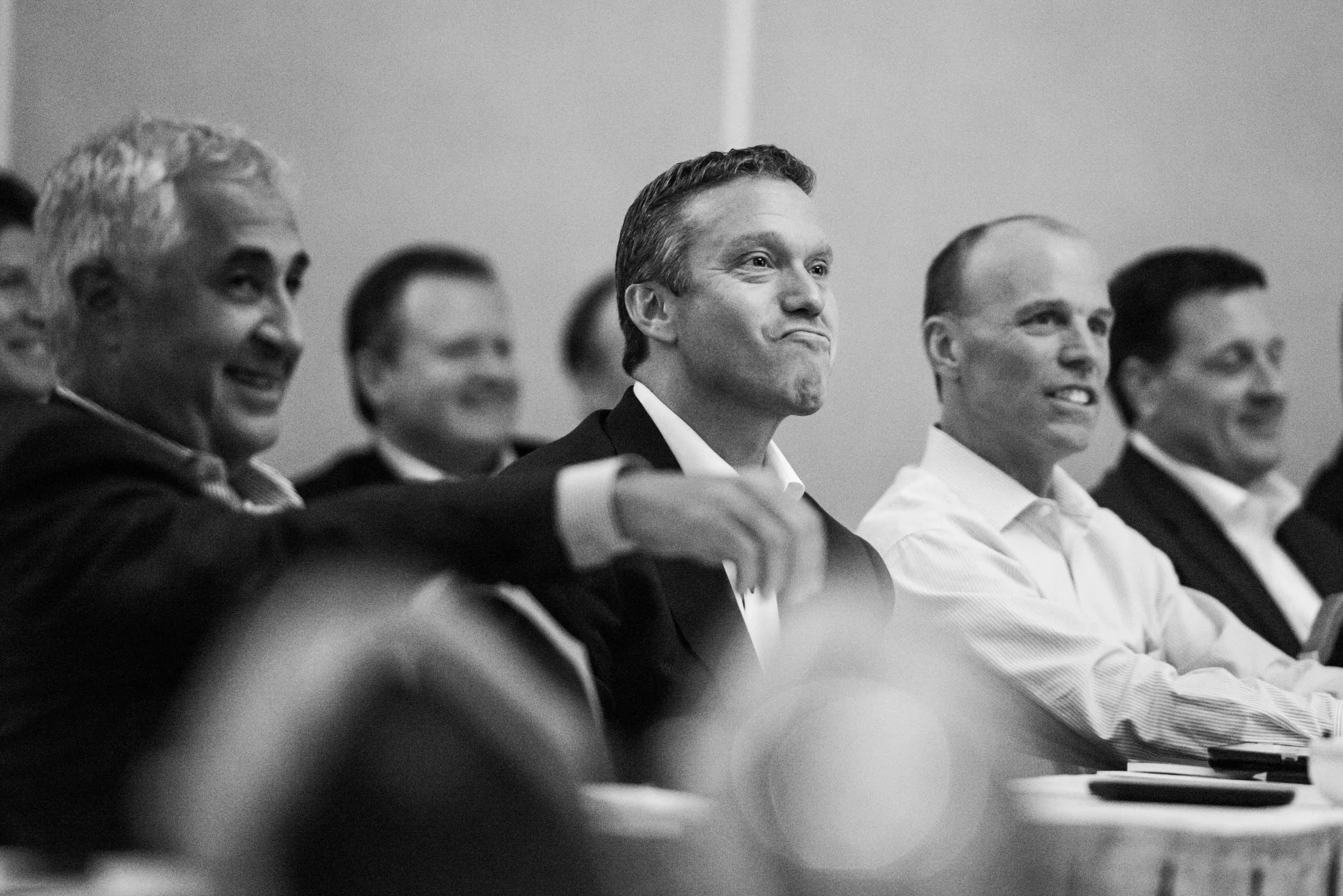 Black and white photo of a group of people in business attire attending a meeting or conference, sitting in a row, smiling and paying attention to something in front of them.