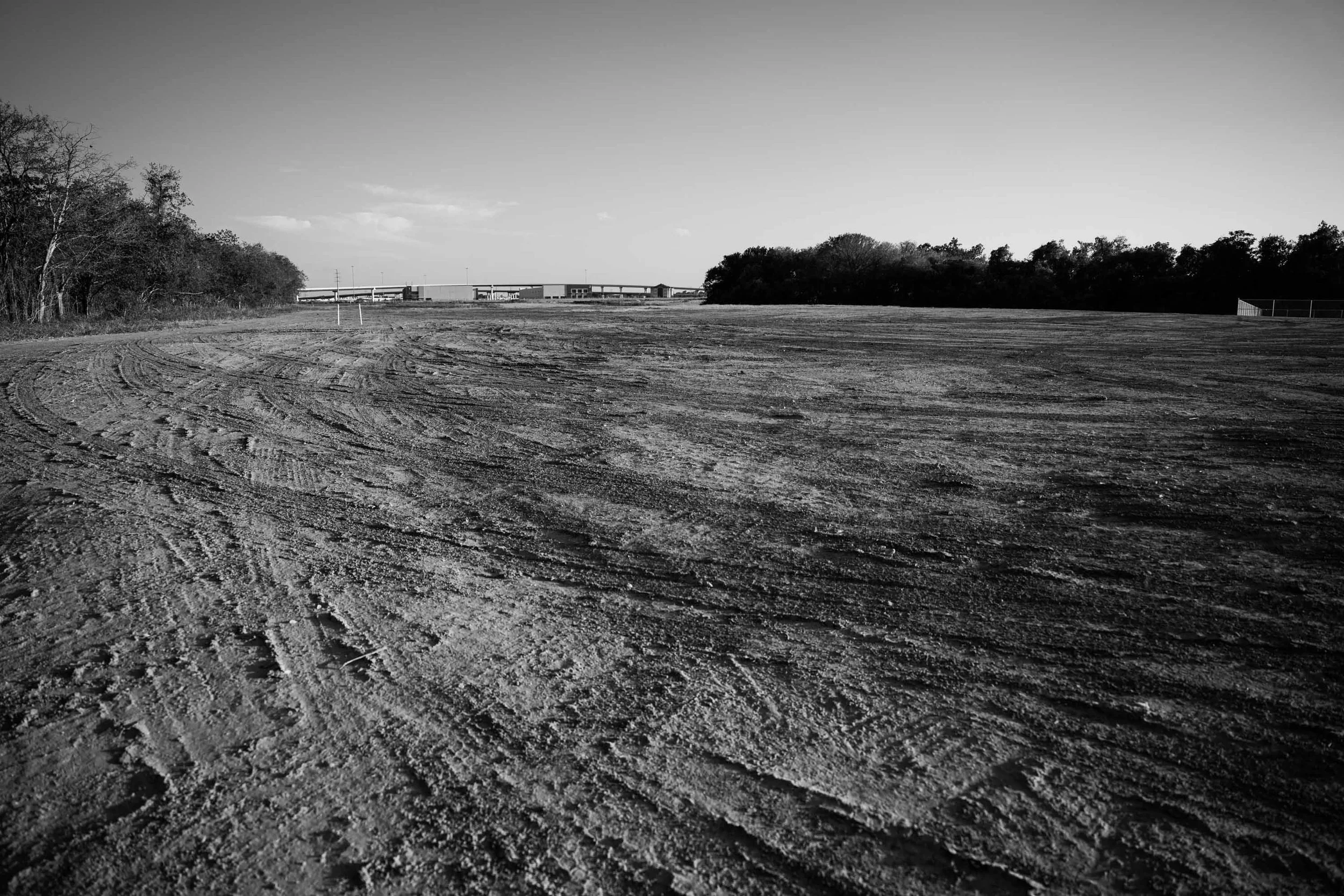 An empty dirt field with tire tracks, with trees on the left and right sides and a bridge or overpass in the distance under a clear sky.