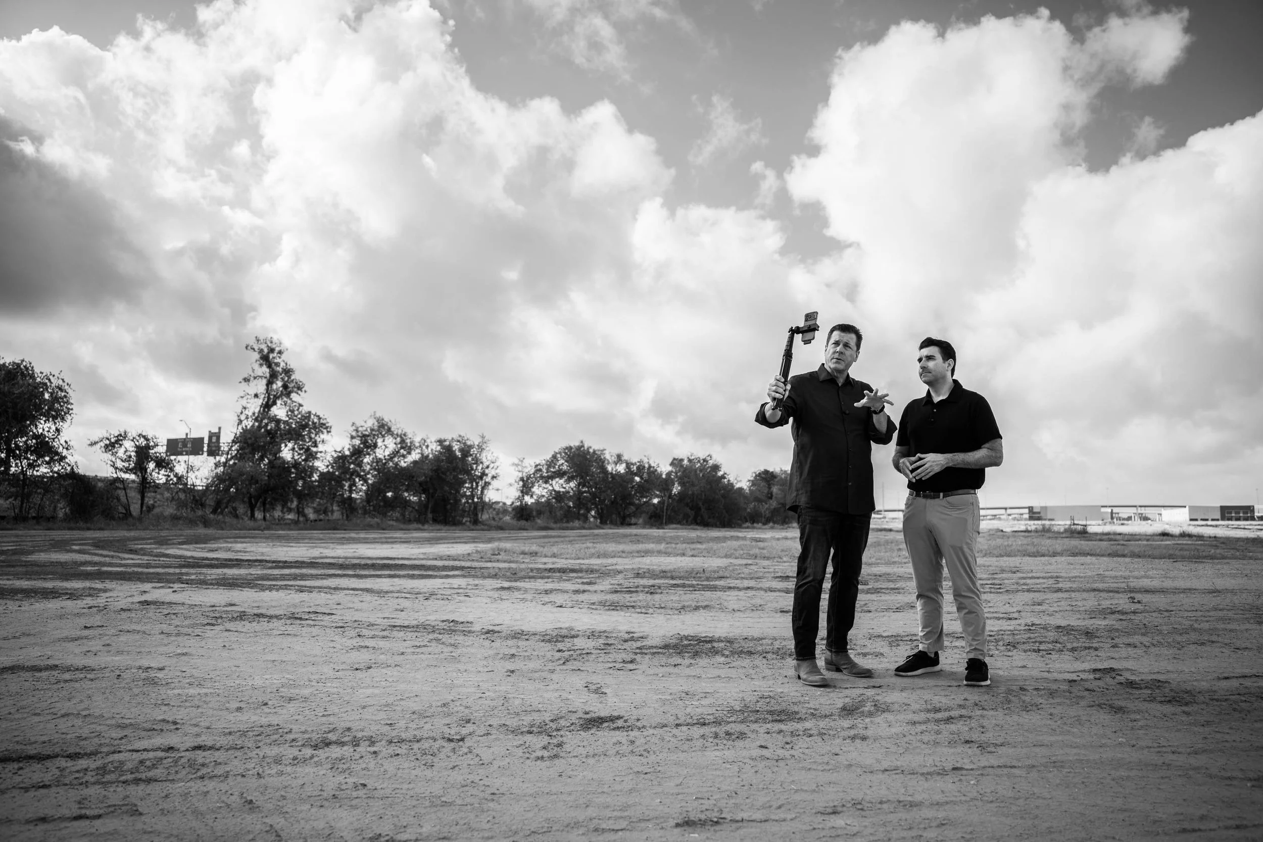 Two men stand outdoors on a dirt field, one holding a selfie stick and the other gesturing with his hand, under a cloudy sky.