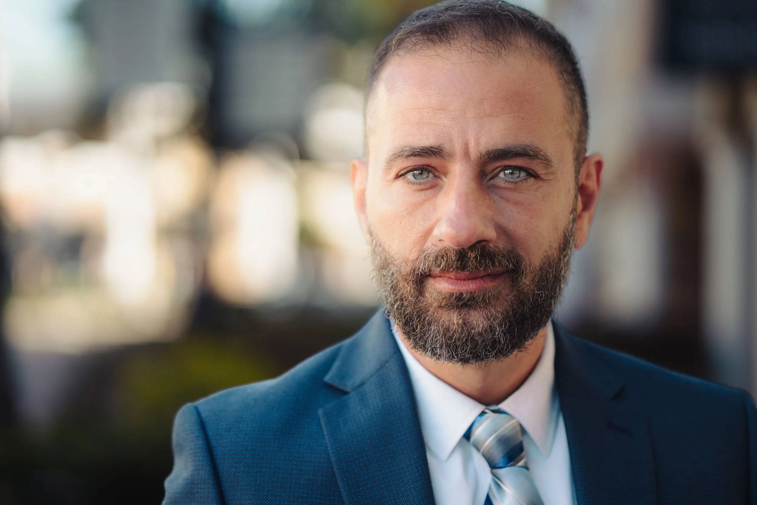 A close-up of a light-skinned man with short brown hair, a full beard, and blue eyes, wearing a dark blue suit, a white shirt, and a diagonally striped light blue and white tie. He is outdoors with a blurred background.