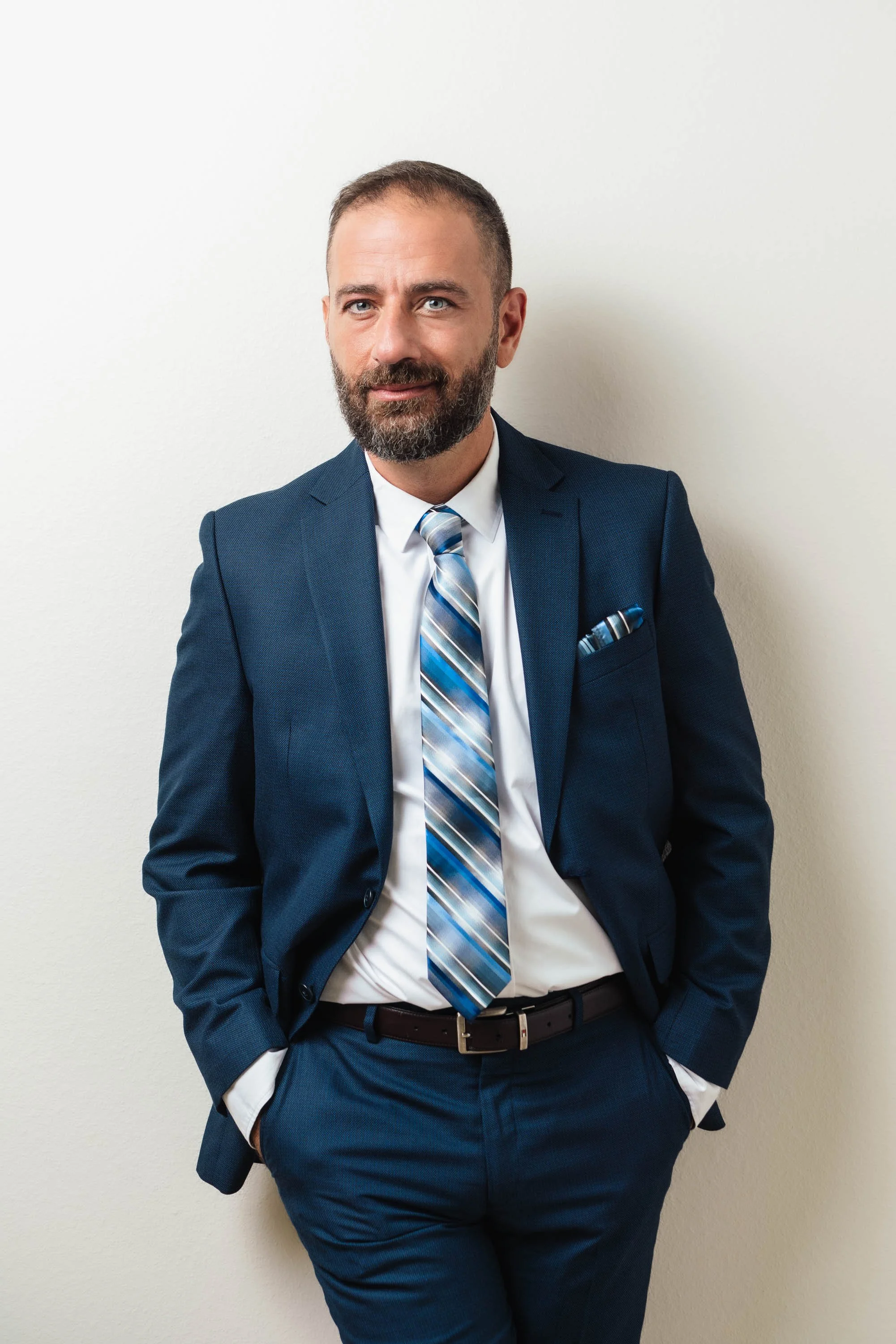 A man with a beard and short hair, dressed in a navy business suit, white shirt, and striped tie, posing with hands in pockets against a plain white wall.