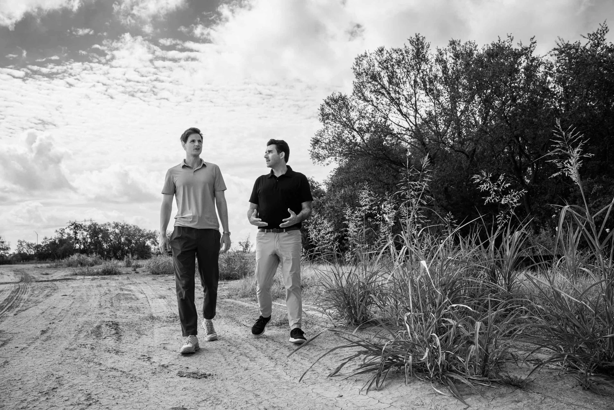 Two men walking and talking on a dirt path outdoors, with trees and bushes in the background, under a partly cloudy sky, in black and white.