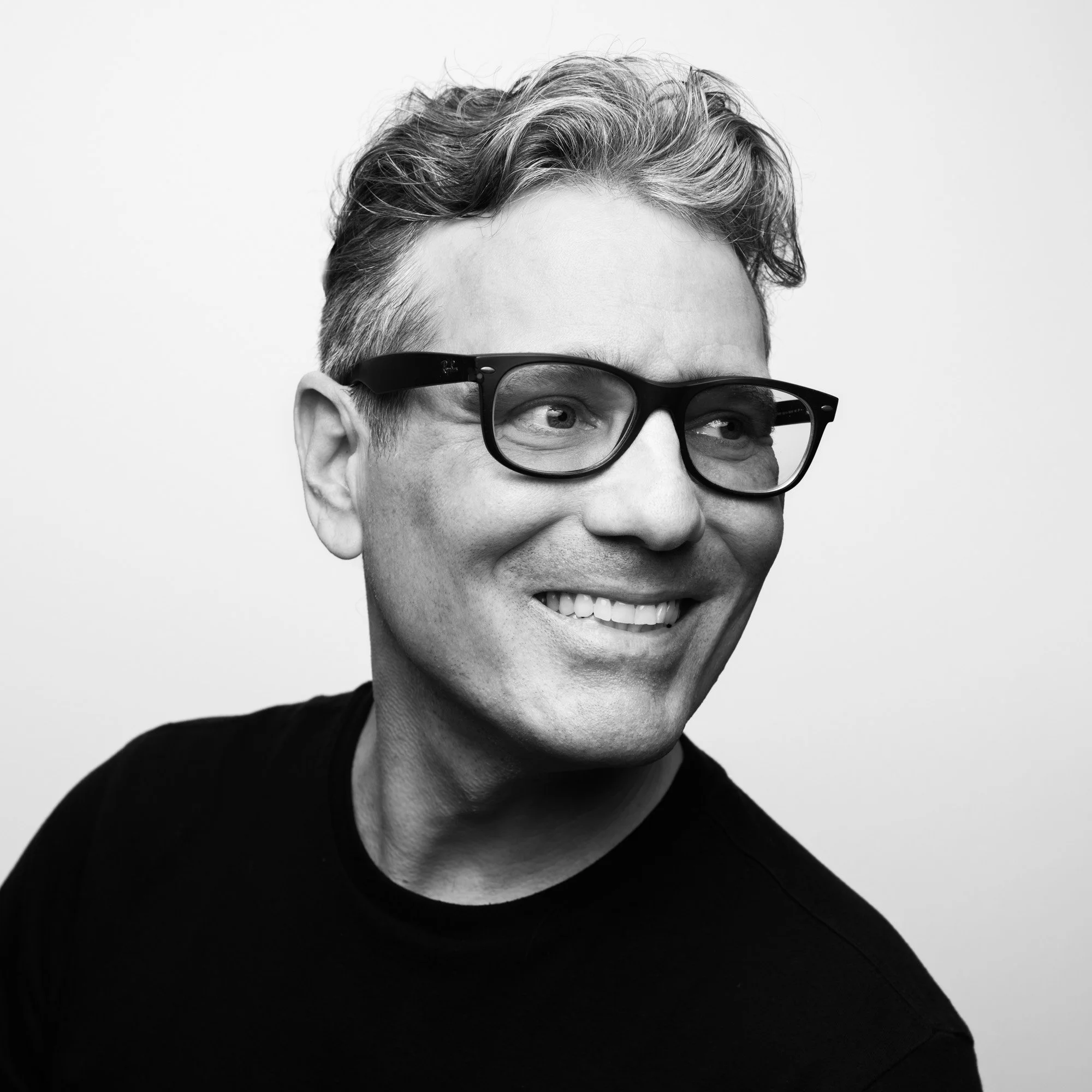 A black-and-white portrait of a smiling man with short, slightly curly hair, wearing black glasses and a dark shirt, against a plain background.