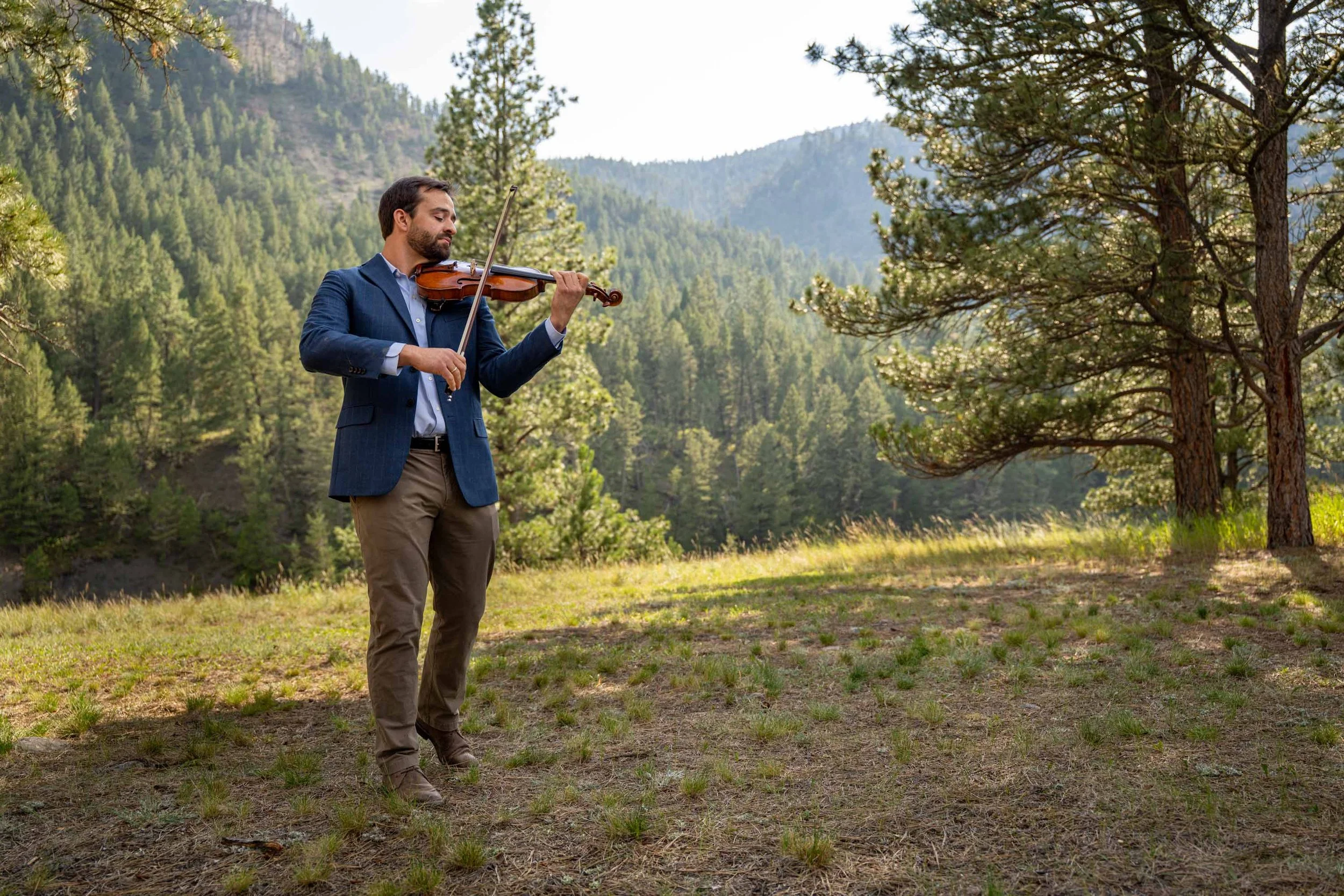 A man in a blue blazer and khaki pants plays the violin outdoors in a forested mountain landscape during daytime.