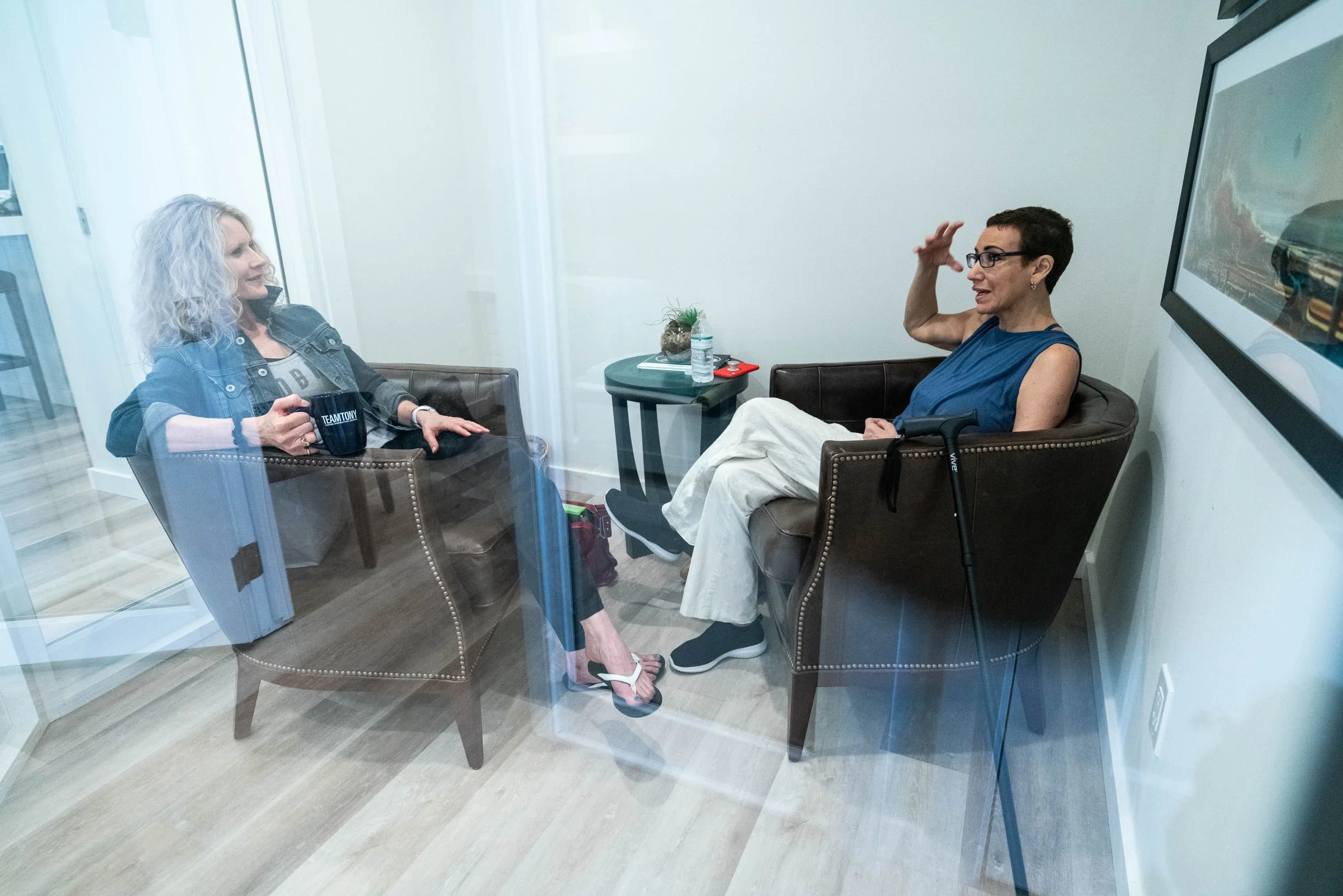 Two women sitting in a cozy office or therapy room, having a conversation through a glass wall, with one woman holding a mug and the other gesturing as she speaks.