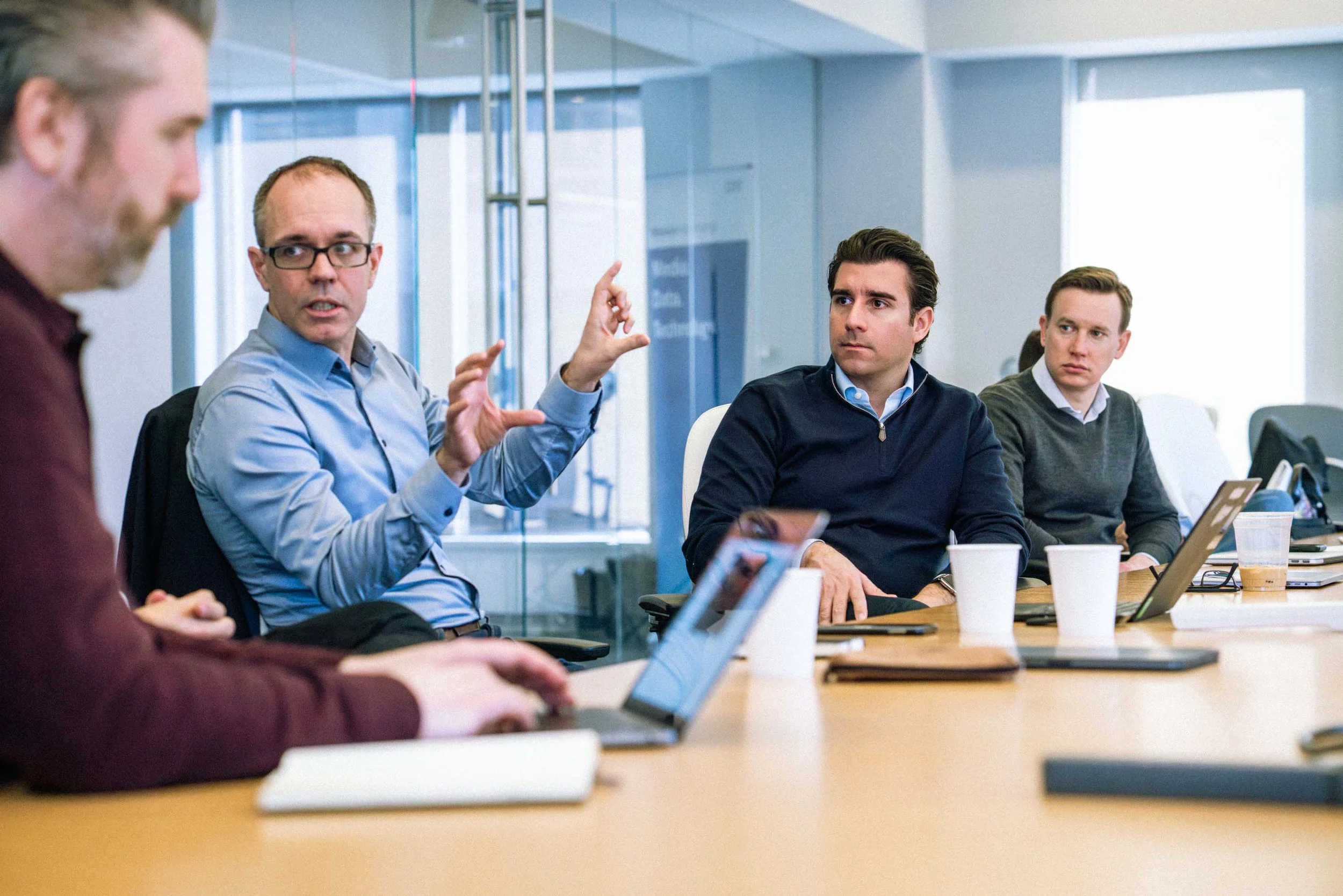 A group of four men sitting at a conference table in a modern office, engaged in a discussion. One man is speaking with hand gestures, while the others listen attentively. There are laptops and coffee cups on the table.