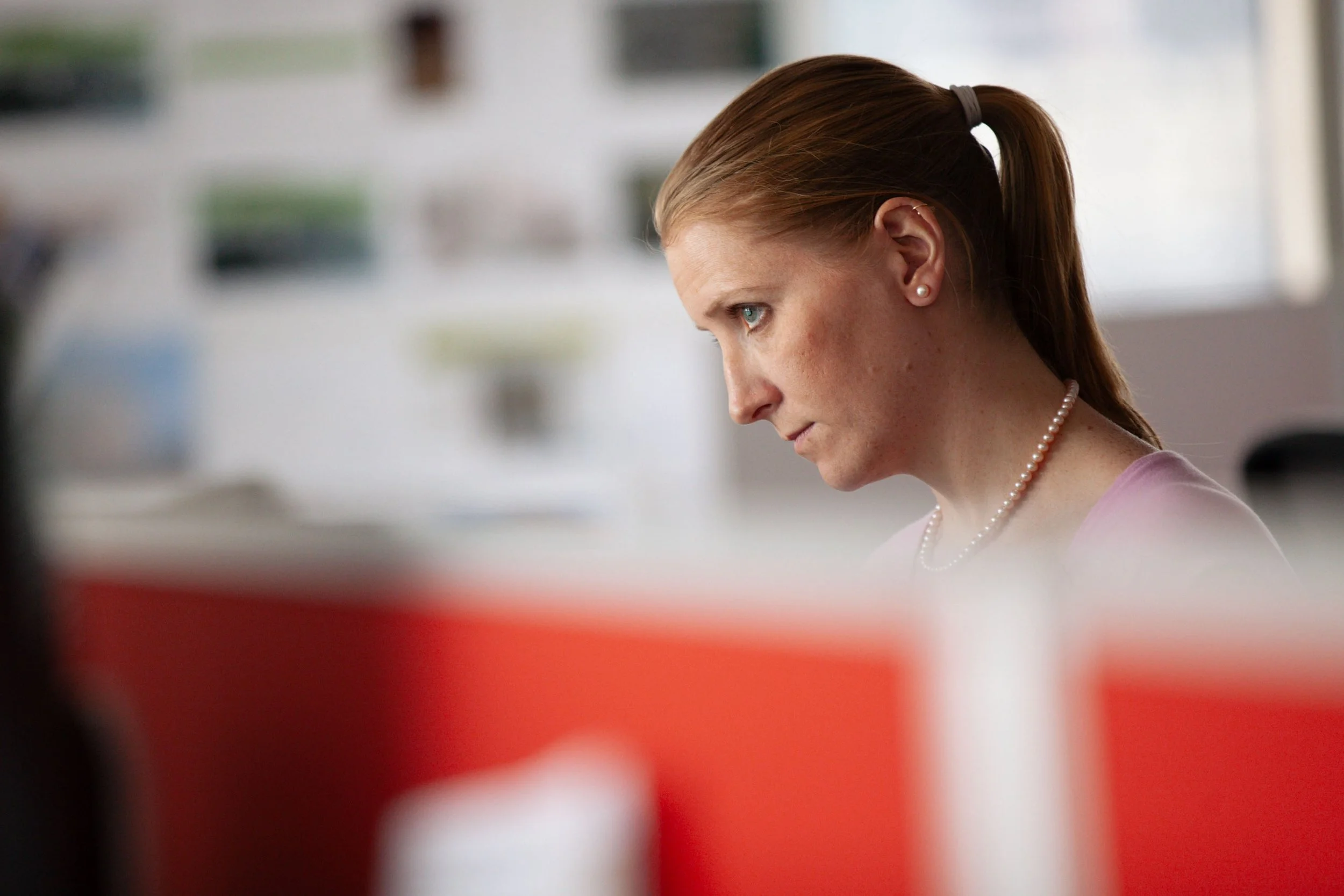 A woman with red hair tied back in a ponytail, wearing pearl earrings and a pearl necklace, is looking down and to her left in an indoor setting.