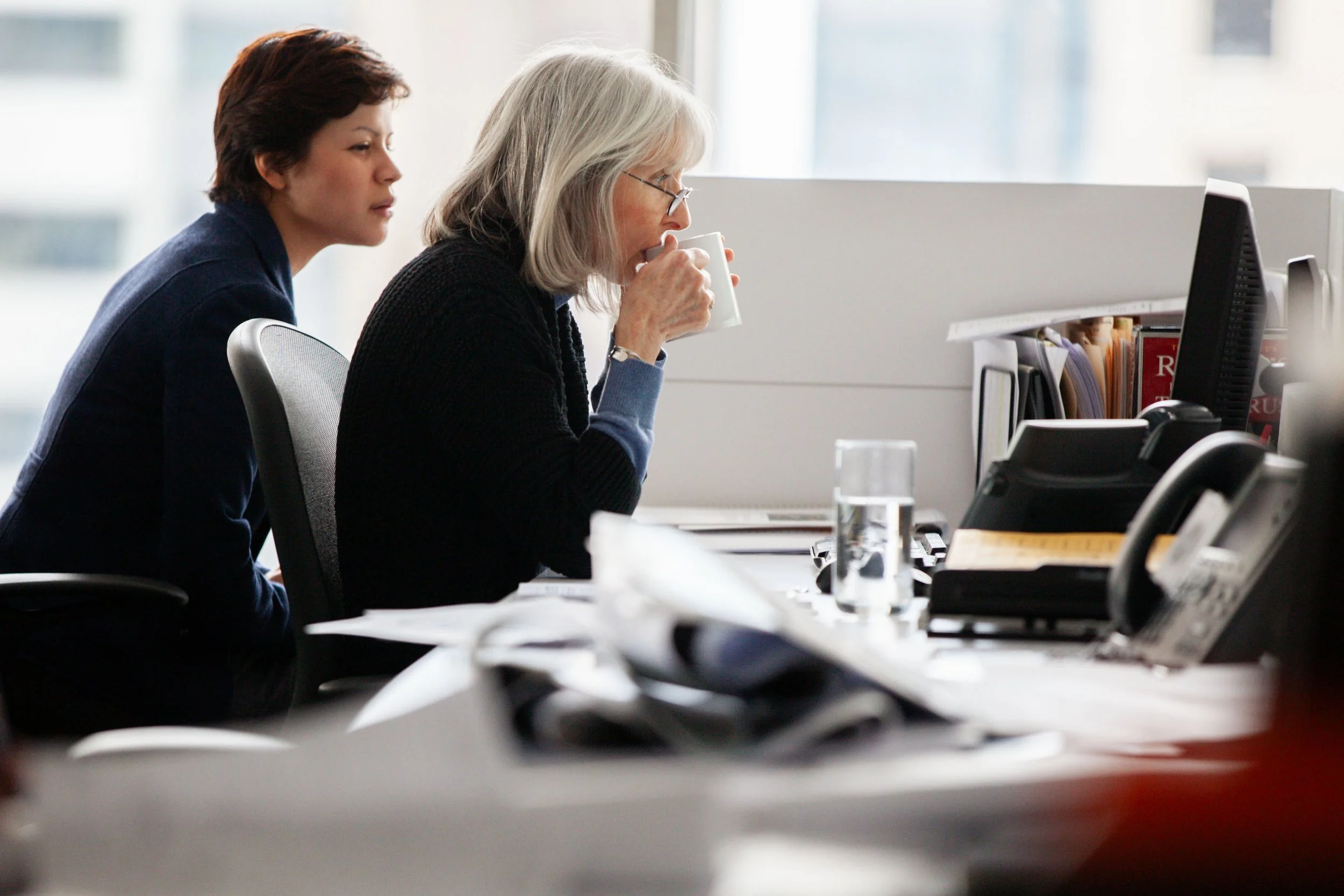 Two women working at desks in an office with computer monitors and office supplies; one woman is drinking coffee, the other is focused on her computer.