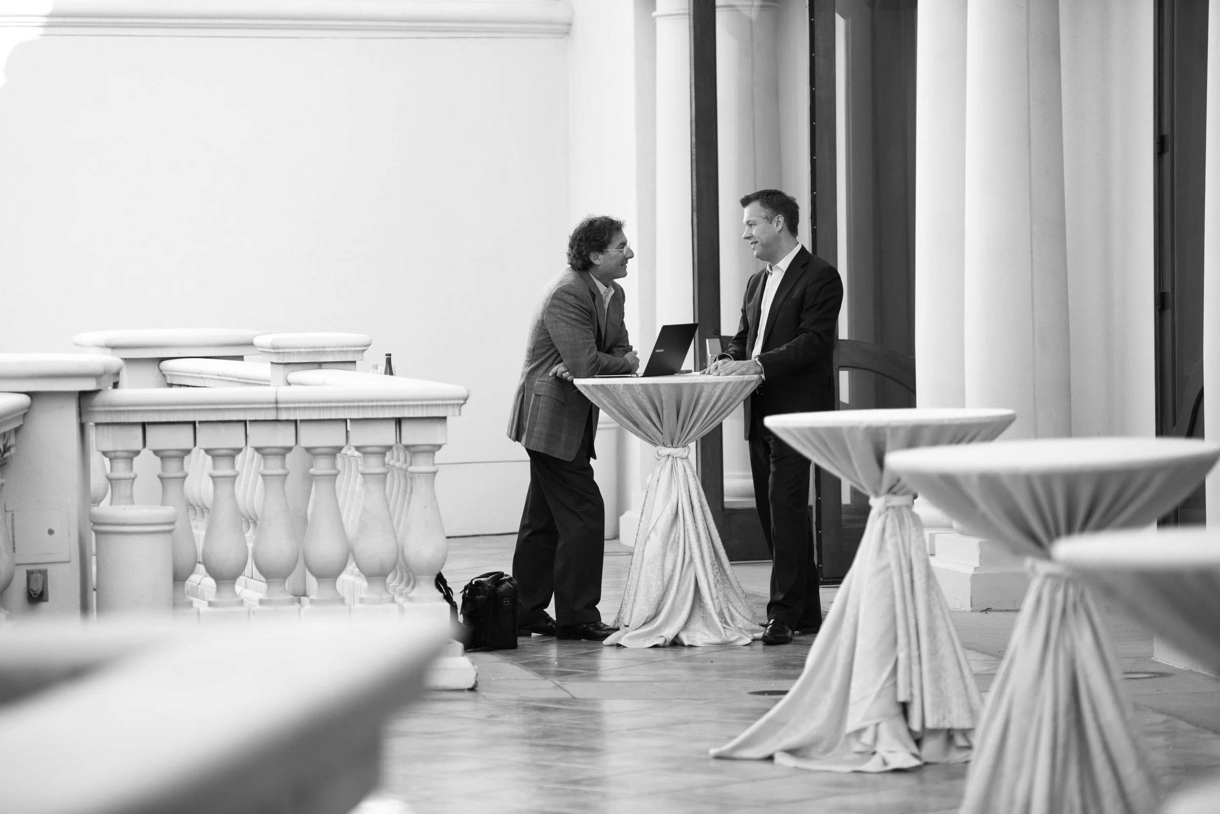 Two men in suits talking at a small round table during a social or professional event, with arranged tables and a large window in the background.