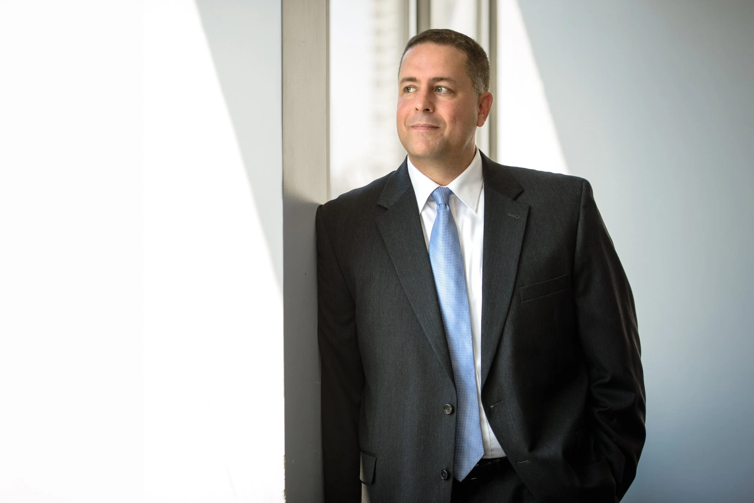 A man in a business suit with a light blue tie, standing by a window, looking out thoughtfully.
