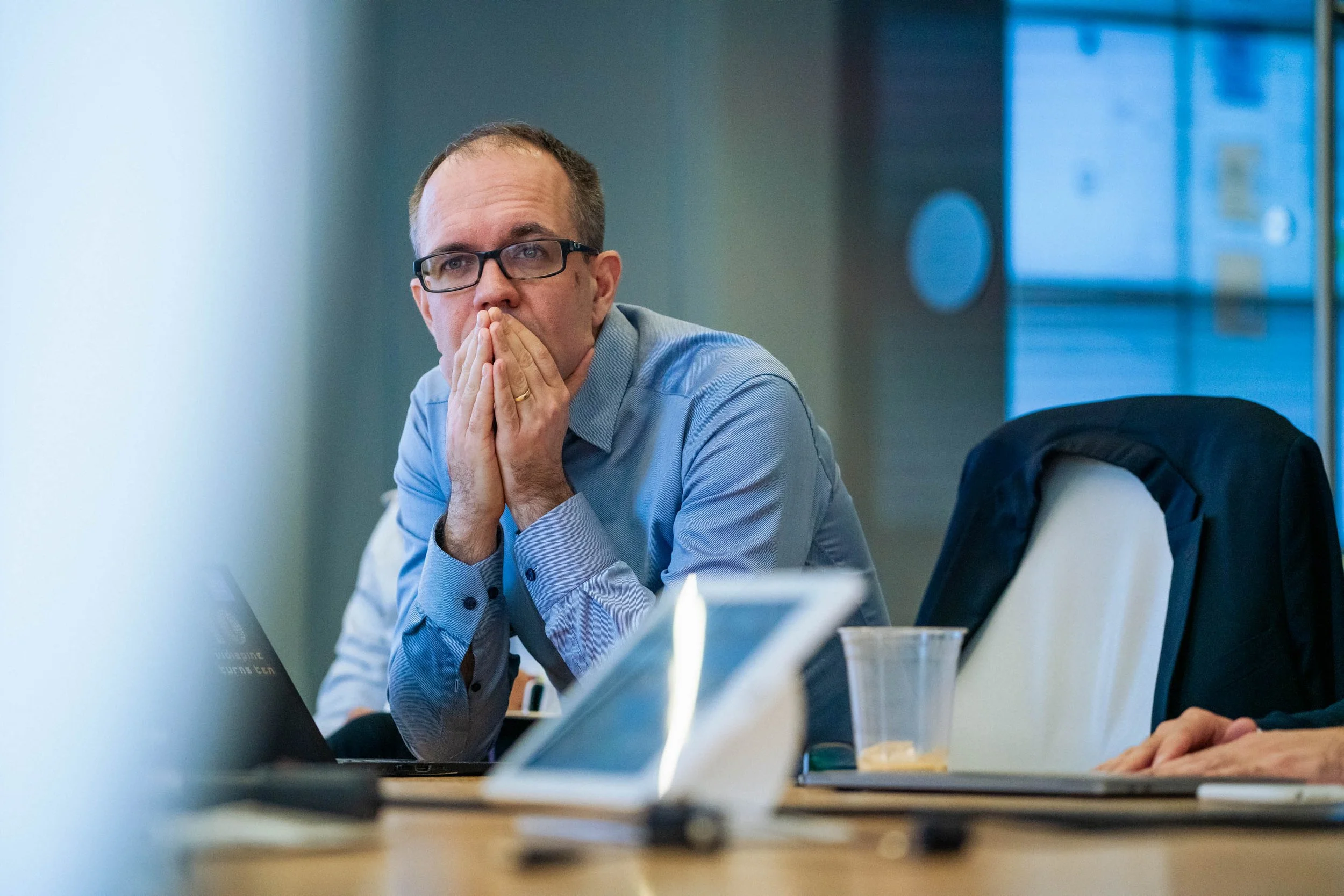 Man wearing glasses with hands covering his mouth, sitting at a table with a laptop and papers.