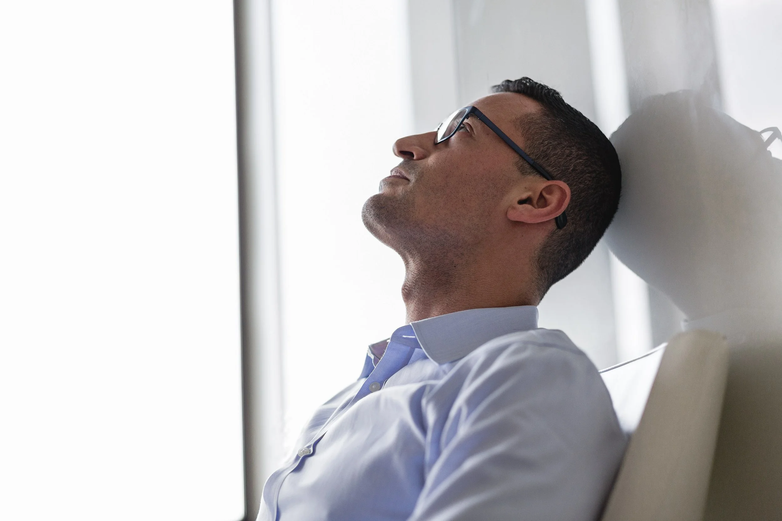 A man with glasses resting his head against a wall, eyes closed, in a light-filled room.