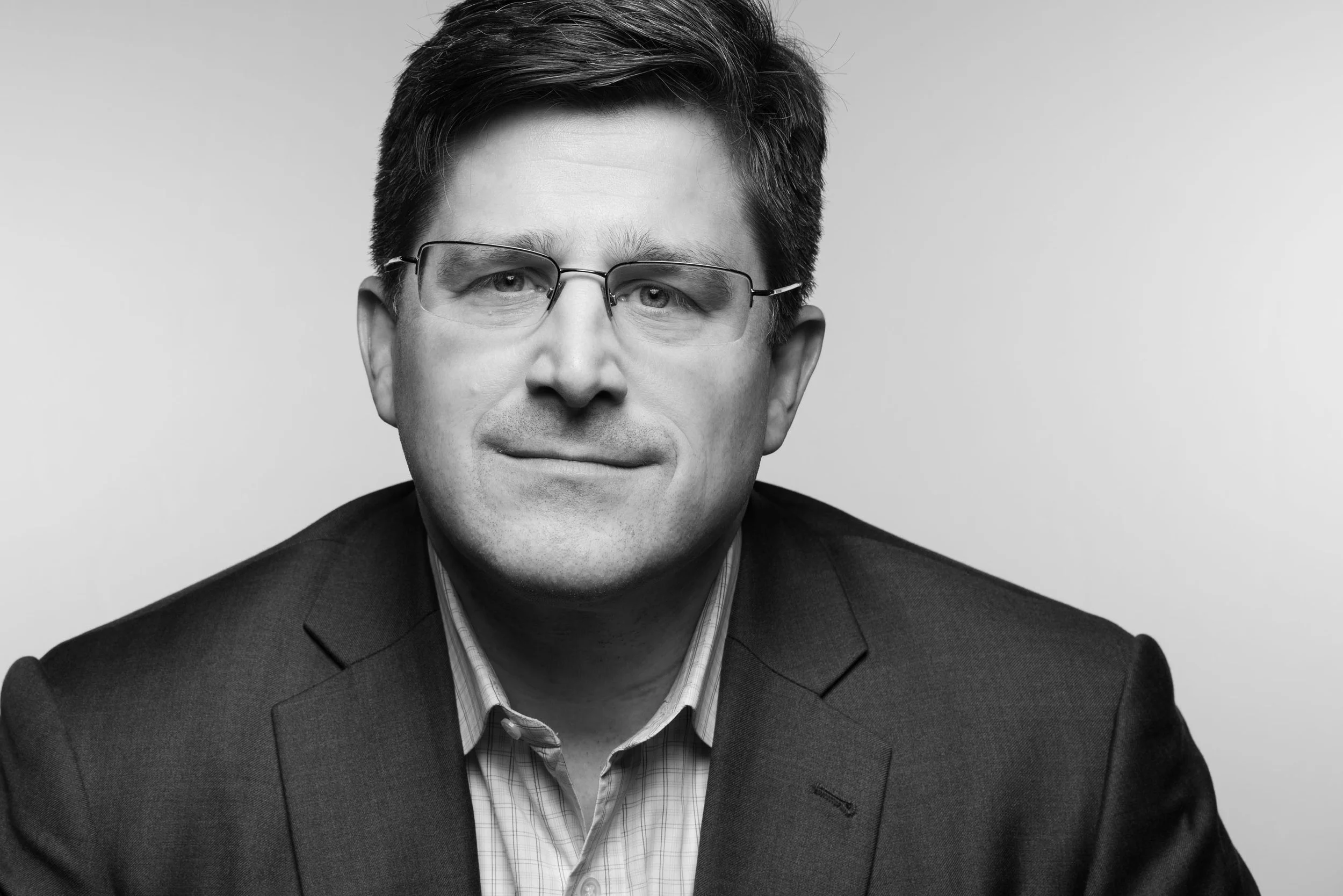 Black-and-white portrait of a man wearing glasses, a plaid shirt, and a suit jacket, smiling slightly against a plain background.
