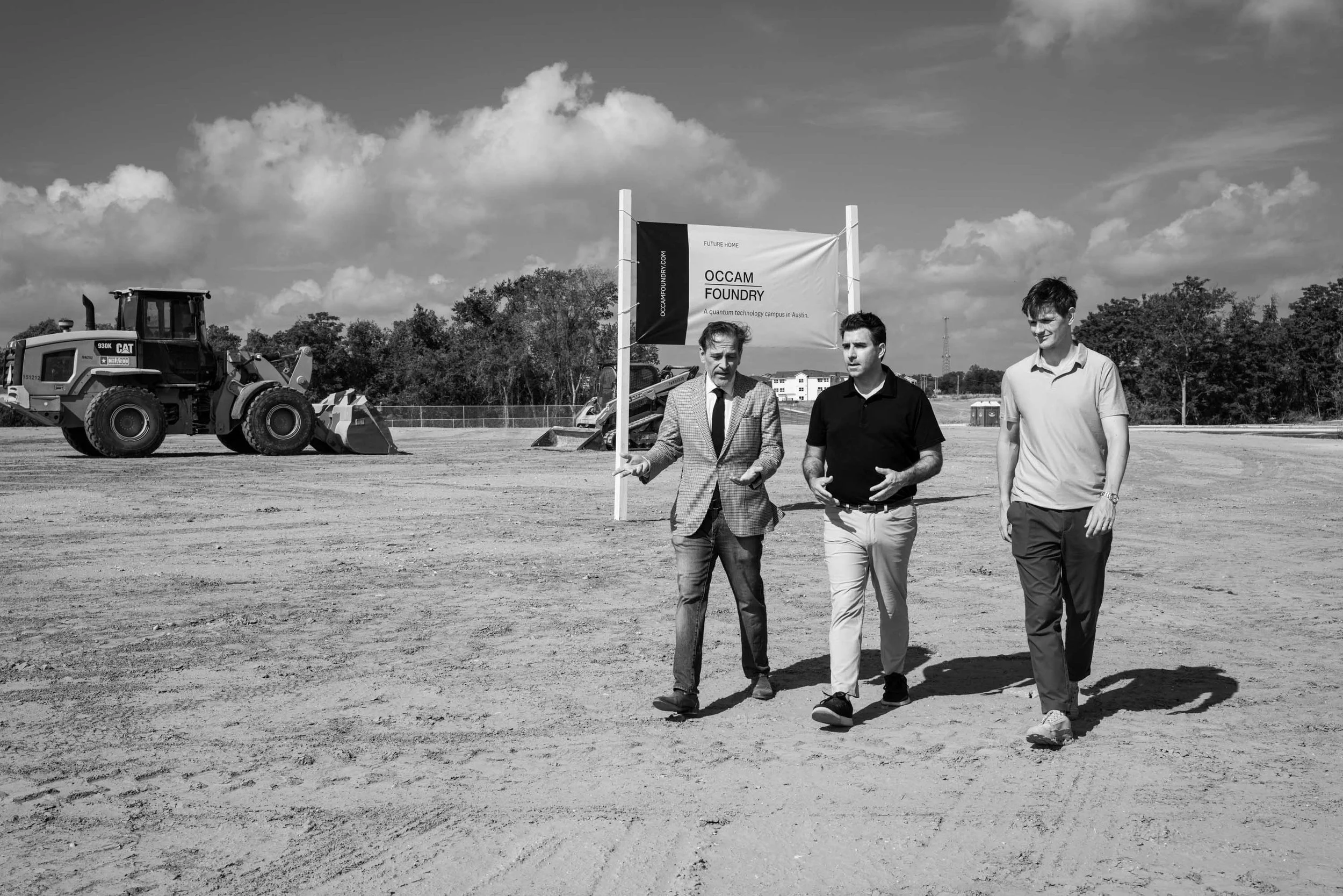 Three men walking and talking on a construction site with a large digger in the background, a sign for OCCAM Foundry, and trees and buildings in the distance.