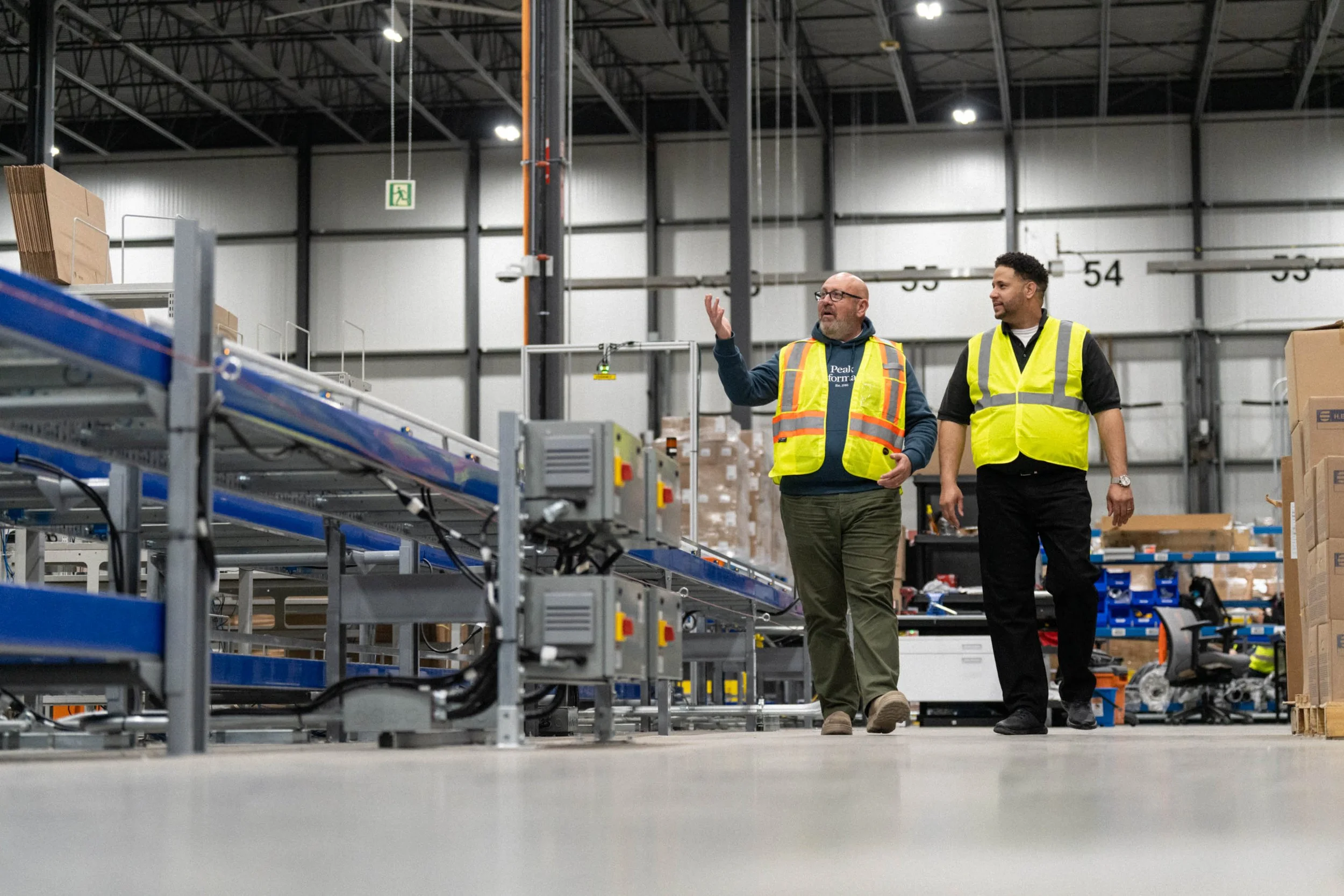 Two men in safety vests walking and talking inside a warehouse or factory with metal shelves and boxes.