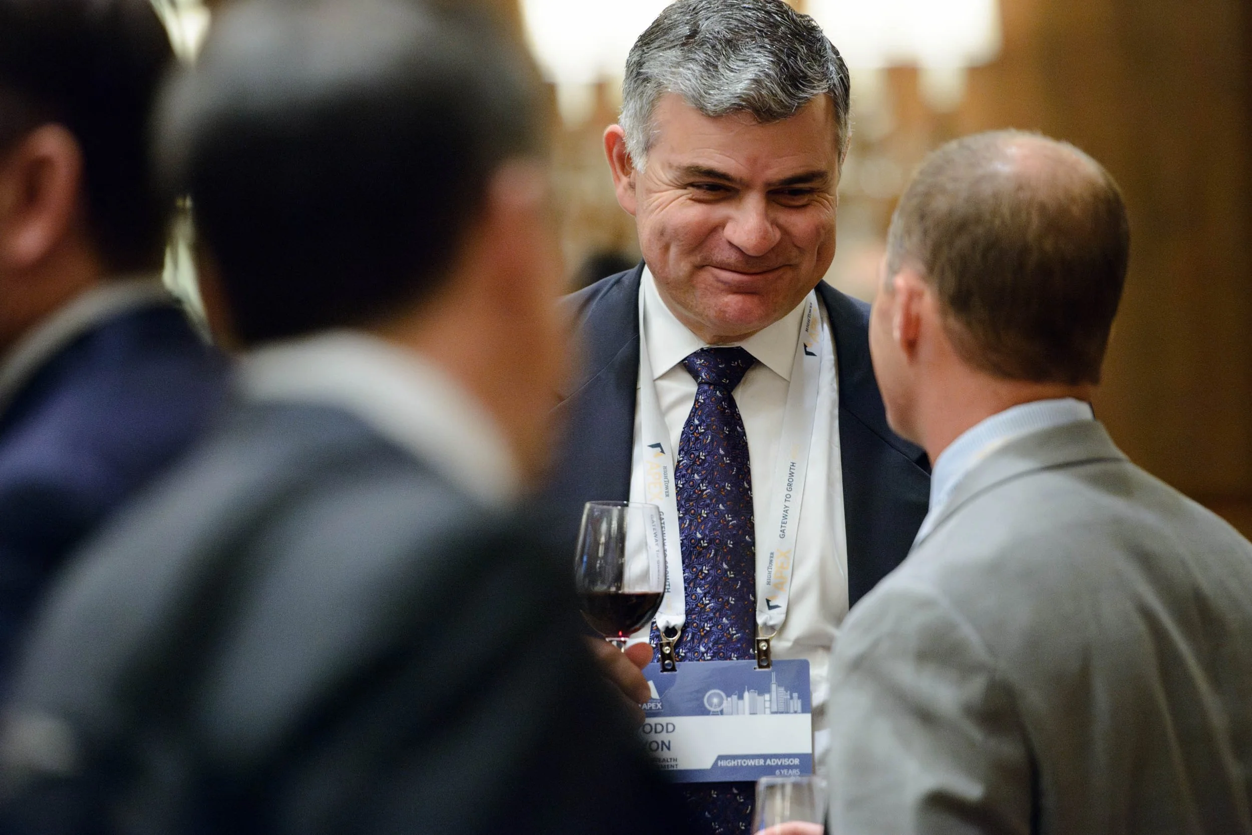 A group of people at a professional event, with one man smiling and holding a glass of red wine, engaged in conversation, wearing a name badge and formal attire.