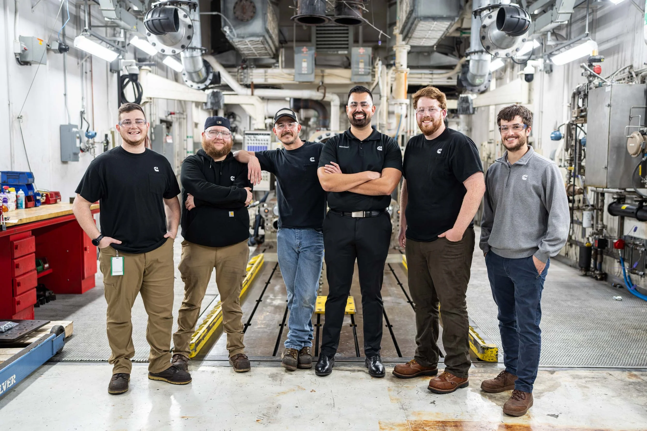 Group of six men standing inside an industrial facility with machinery and pipes in the background.