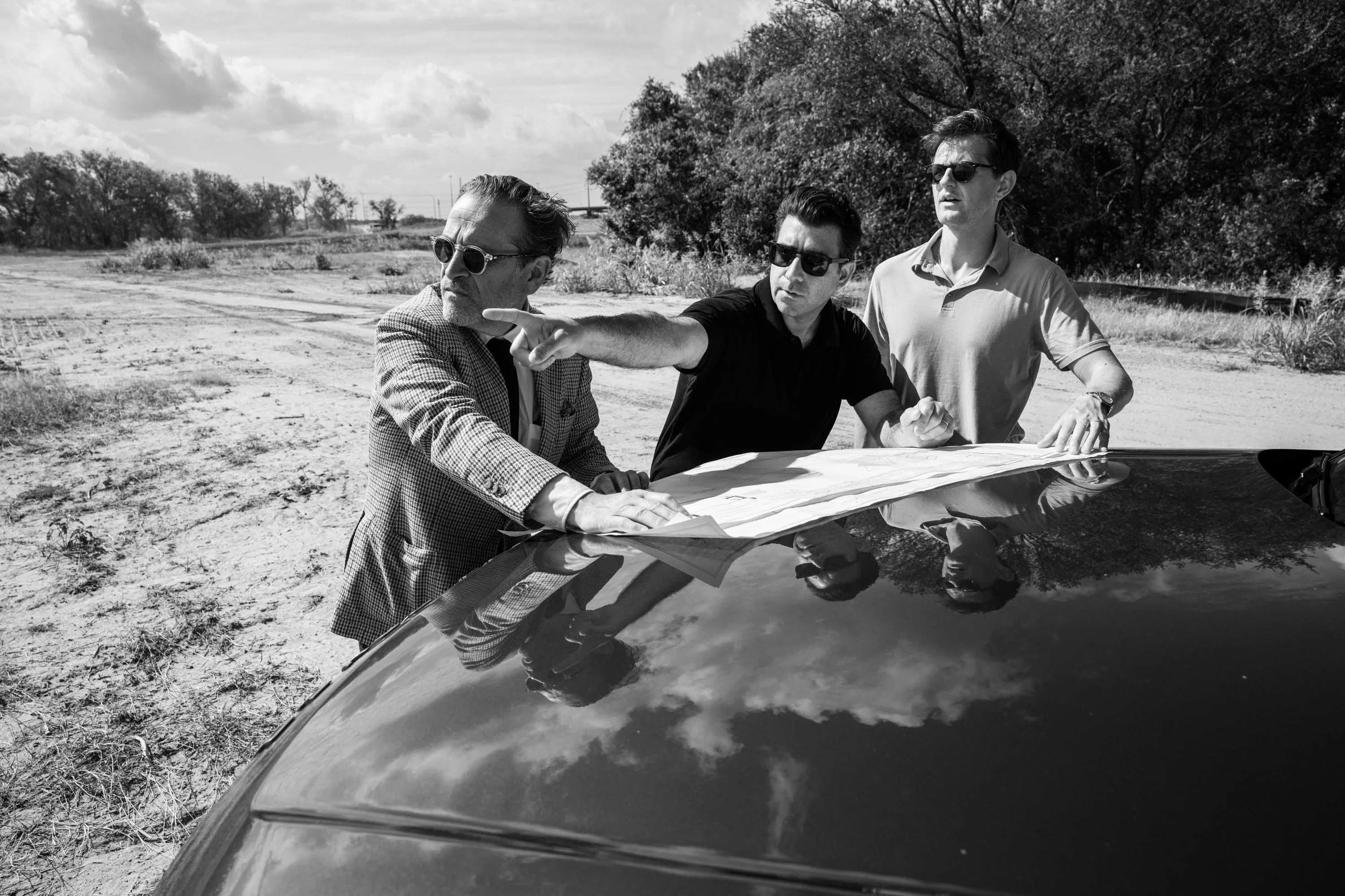 Three men standing outdoors on a dirt path near trees, looking at a map on the hood of a car, with one pointing, all wearing sunglasses.