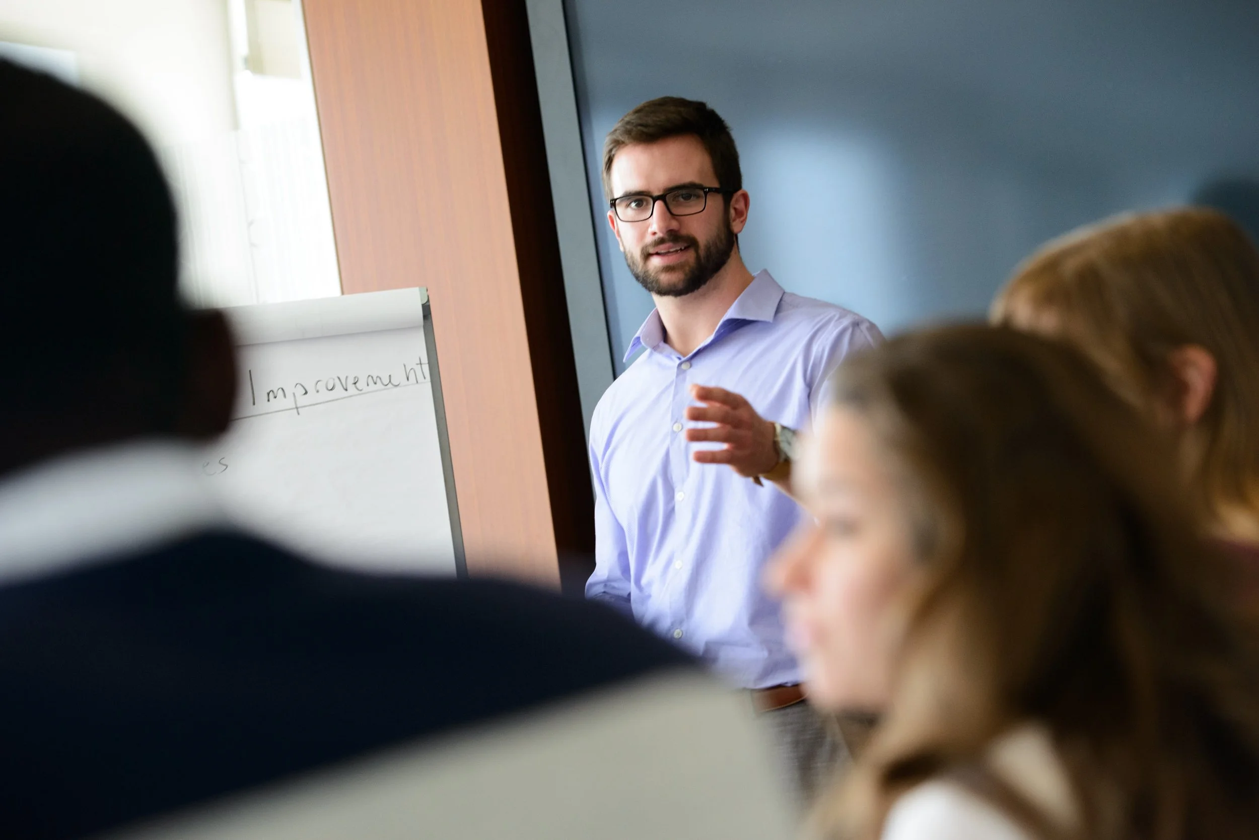 A man with glasses and a beard is giving a presentation in front of a classroom, with students listening. The whiteboard behind him has the word 'improvement' partially visible.