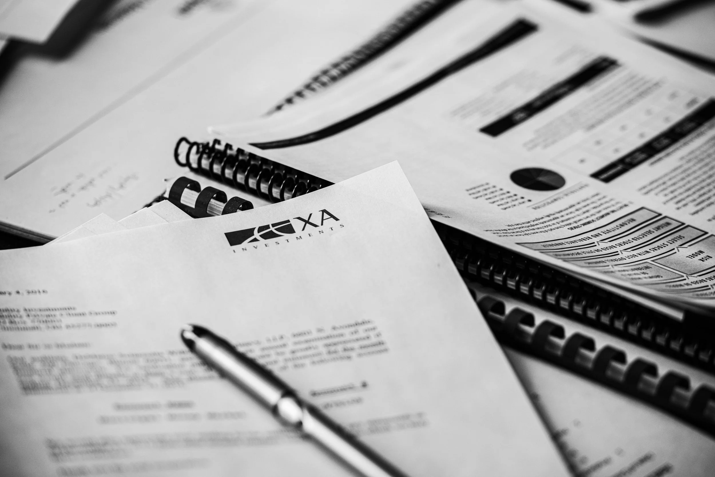 Black and white photo of various documents and financial papers, with a logo labeled 'AXA Investments,' along with spiral-bound notebooks and a pen.