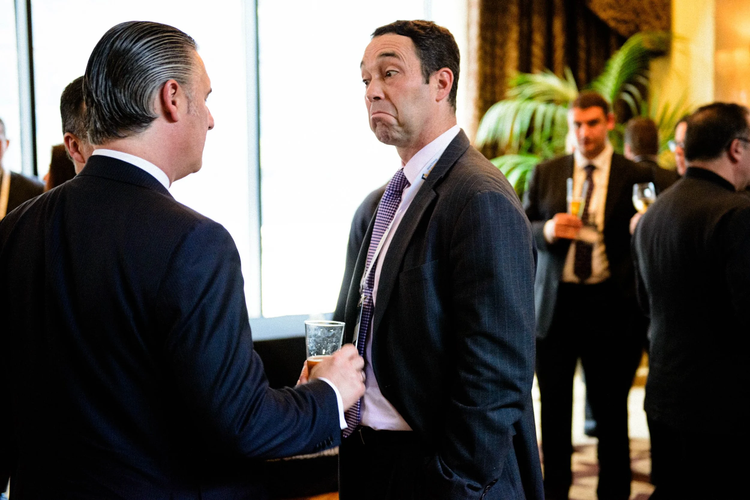 Two men in suits engaging in conversation at a social event, one holding a pint of beer, with others mingling in the background.