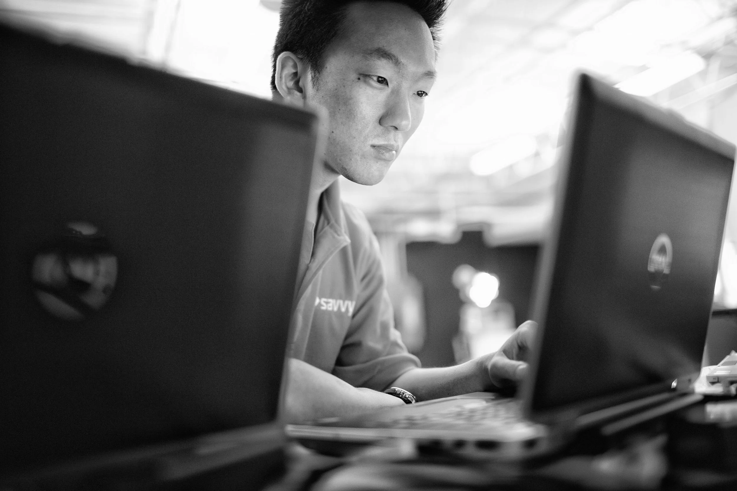 A young man working on a laptop in a bright, indoor setting, with other laptops visible in the foreground.