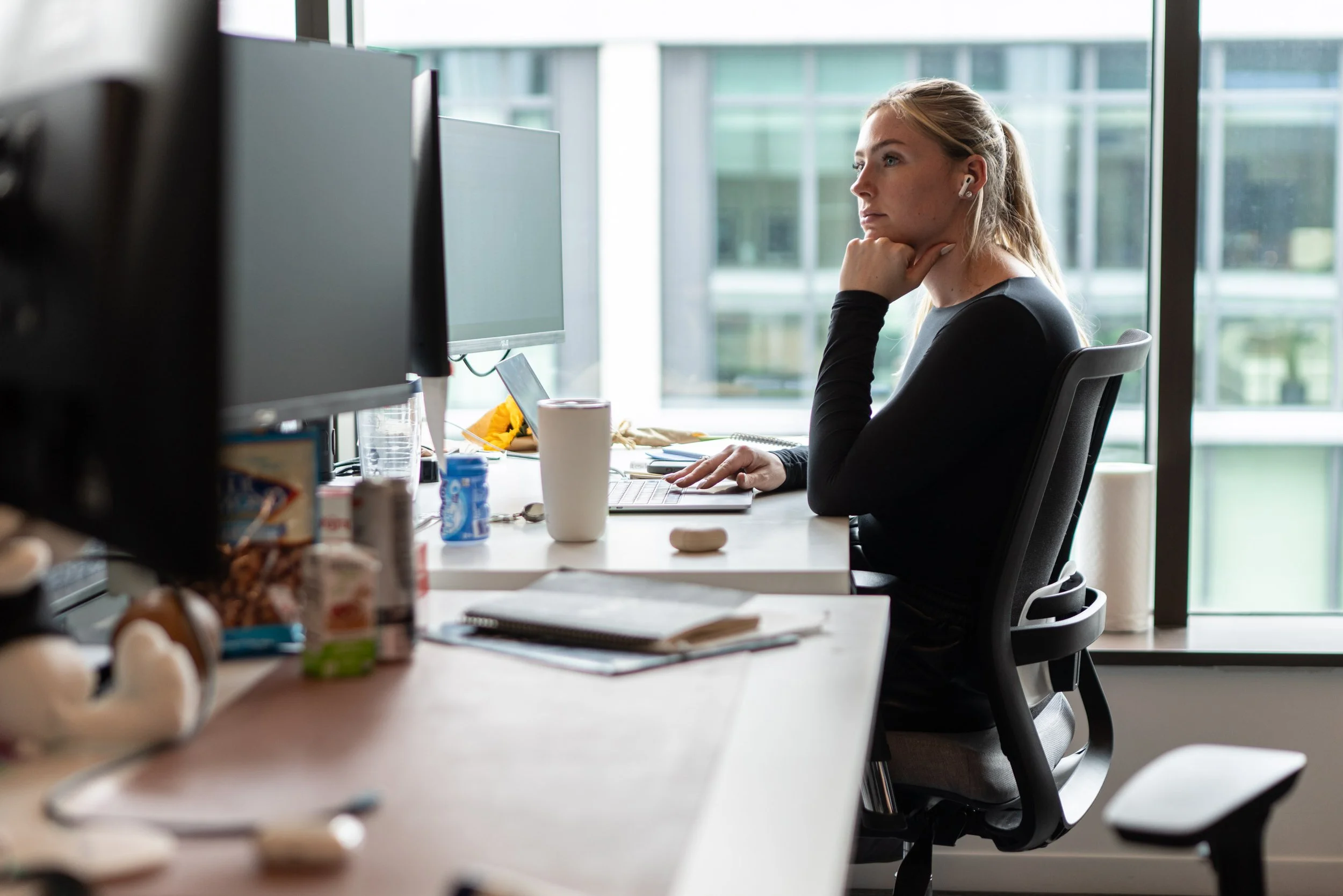 A woman working at a desk with multiple computer monitors in a modern office, with a glass window showing a cityscape outside.