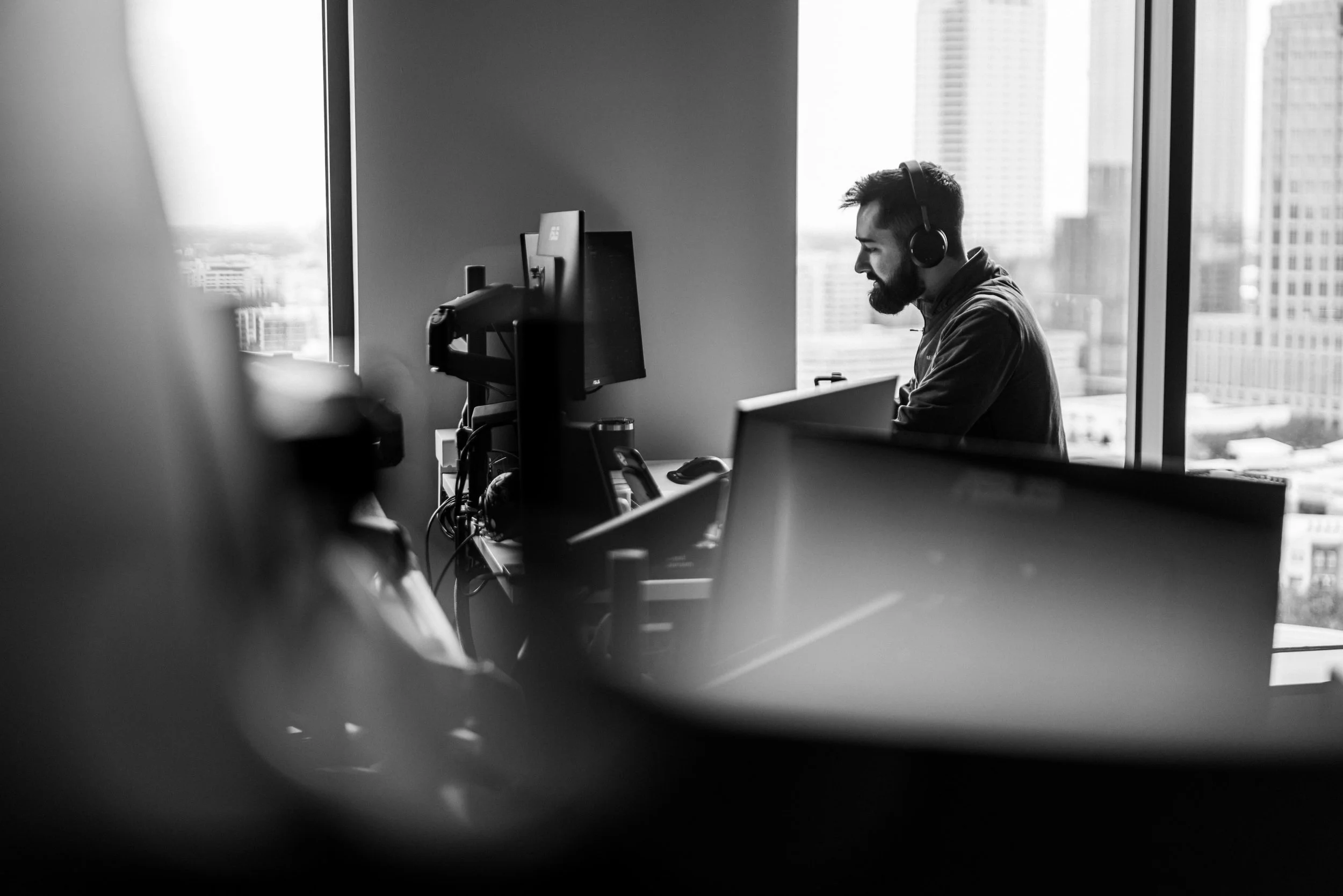 A man with a beard wearing headphones working at a desk with a computer, in a high-rise office with large windows showing a city view, black and white photo.