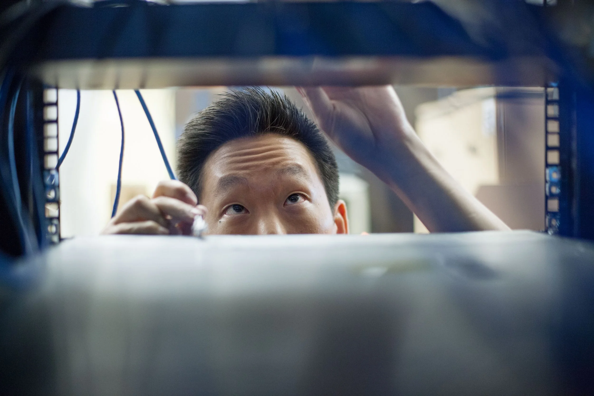 A person with neatly styled dark hair and light skin is seen through a small opening, looking up while holding something in their right hand. The perspective appears to be from below, with shelves and cables visible around the person.
