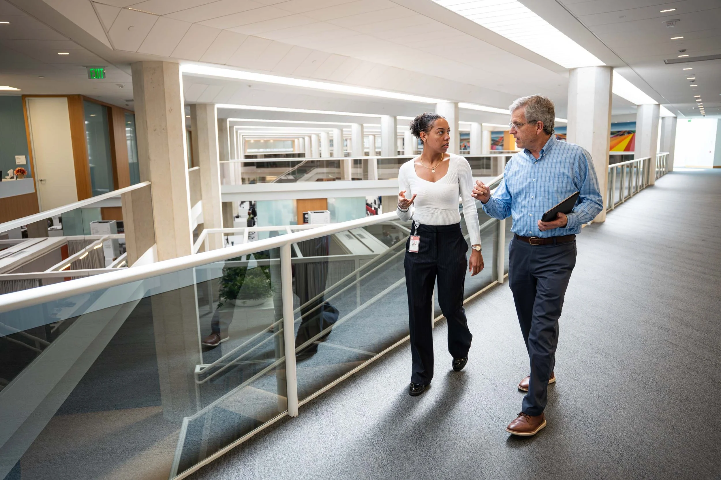 Two professionals, a woman and a man, are walking and talking in a modern office building hallway with glass railings and bright lighting.