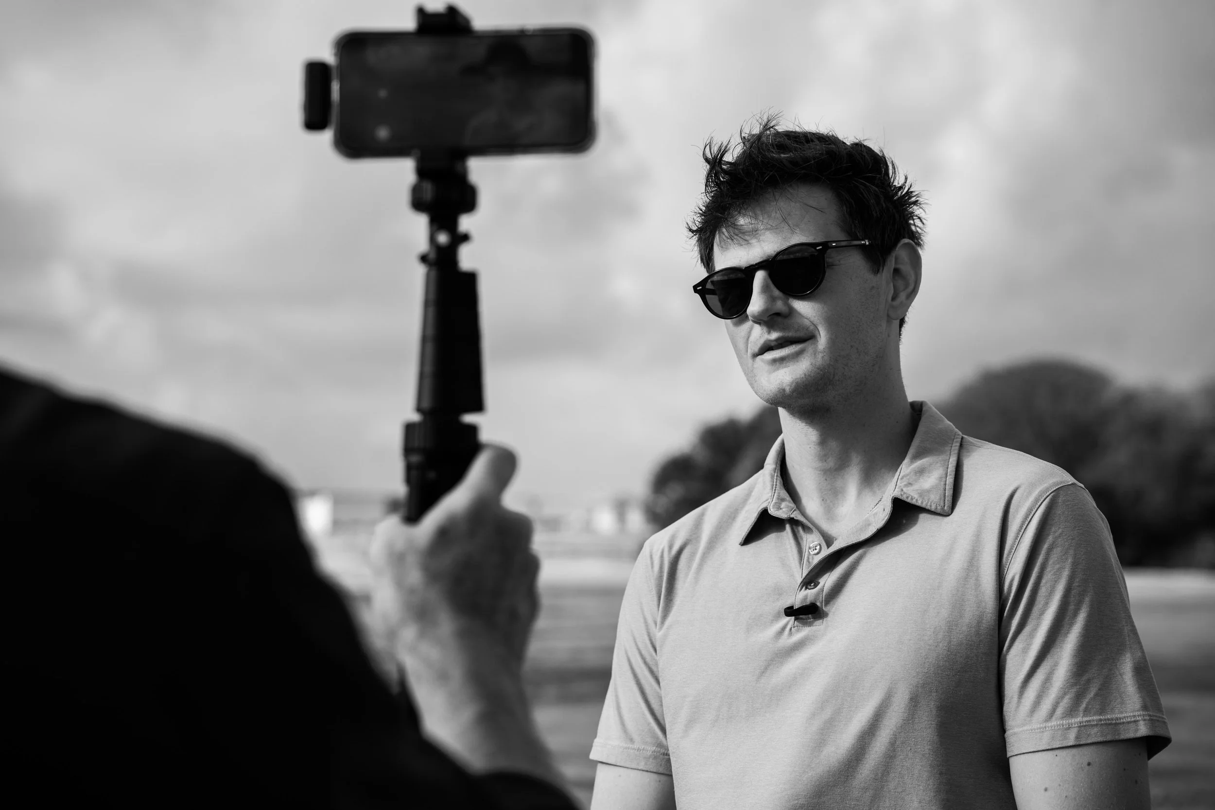 A young man wearing sunglasses is being recorded by another person using a smartphone mounted on a selfie stick outdoors, with a cloudy sky in the background.