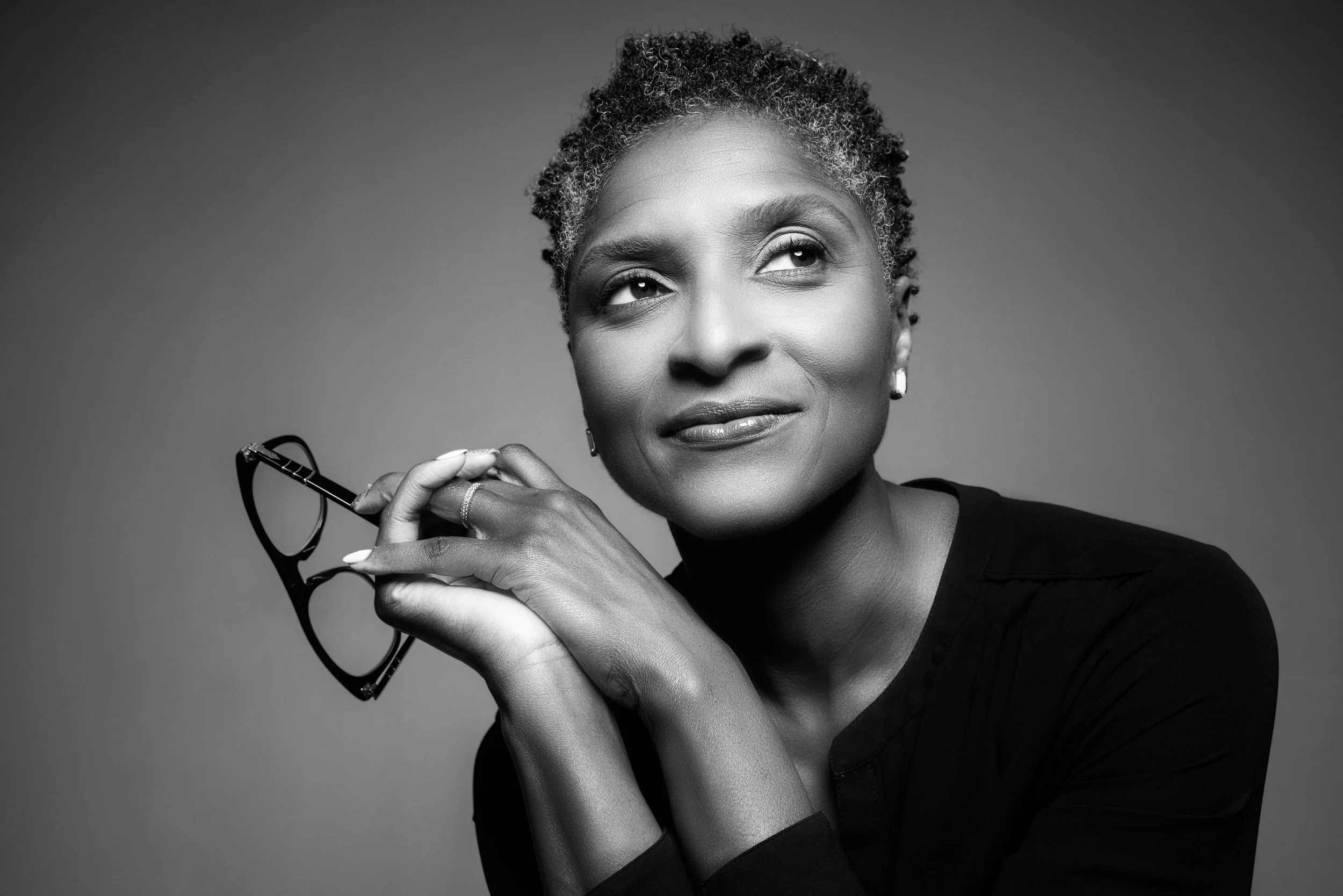 Black and white portrait of an elegant woman with short curly hair, holding glasses with one hand near her face, wearing earrings and a dark top, against a plain background.