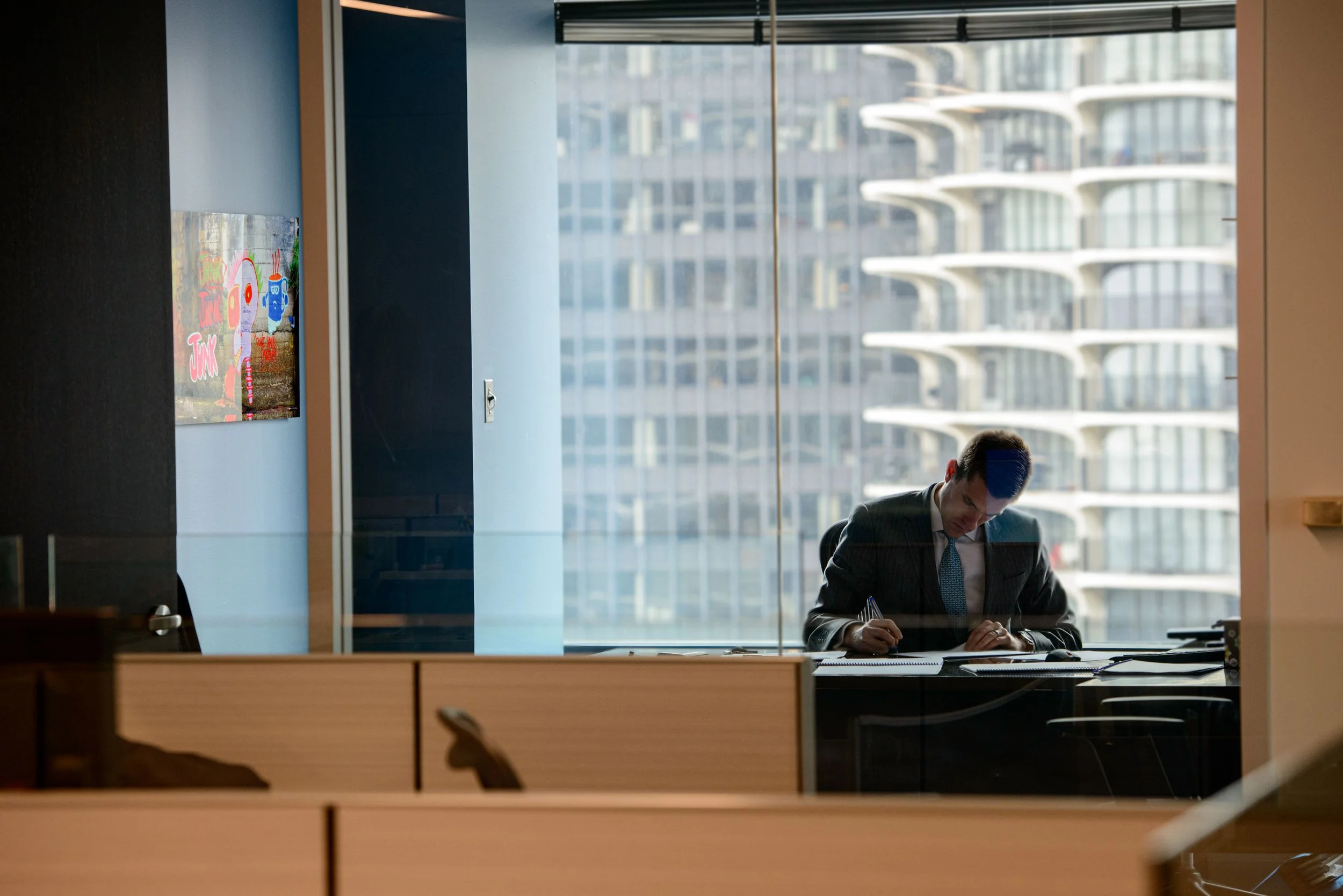 A man in a business suit and tie sitting at a desk in a modern office with large windows showing high-rise buildings outside, working and writing.