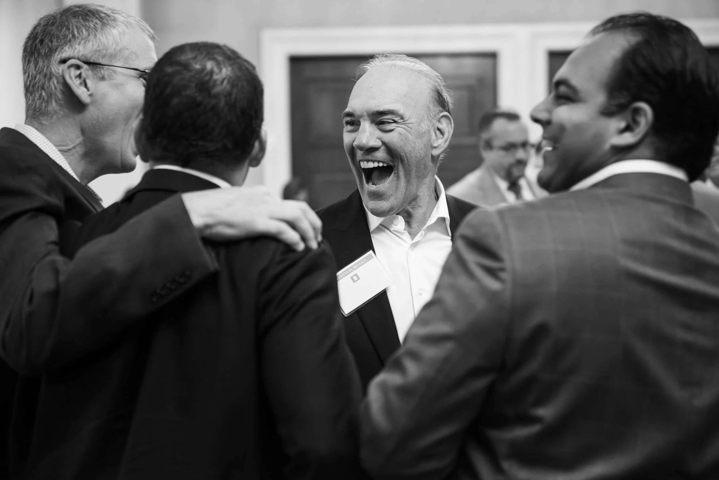 Smiling man with a name badge engaging in conversation with other men in formal attire at a professional event.