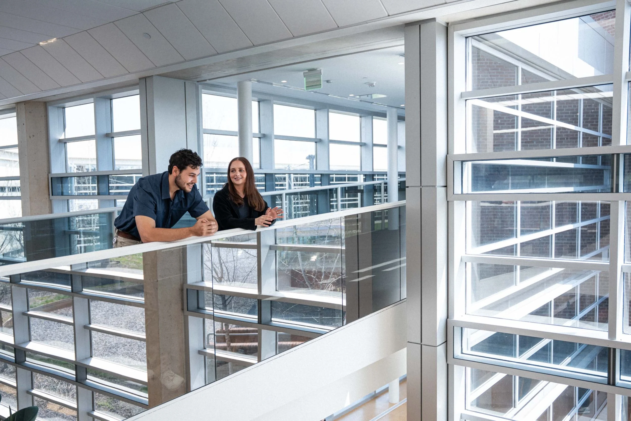 Two people, a man and a woman, are leaning on a railing and talking inside a modern building with large windows and multiple stories visible.