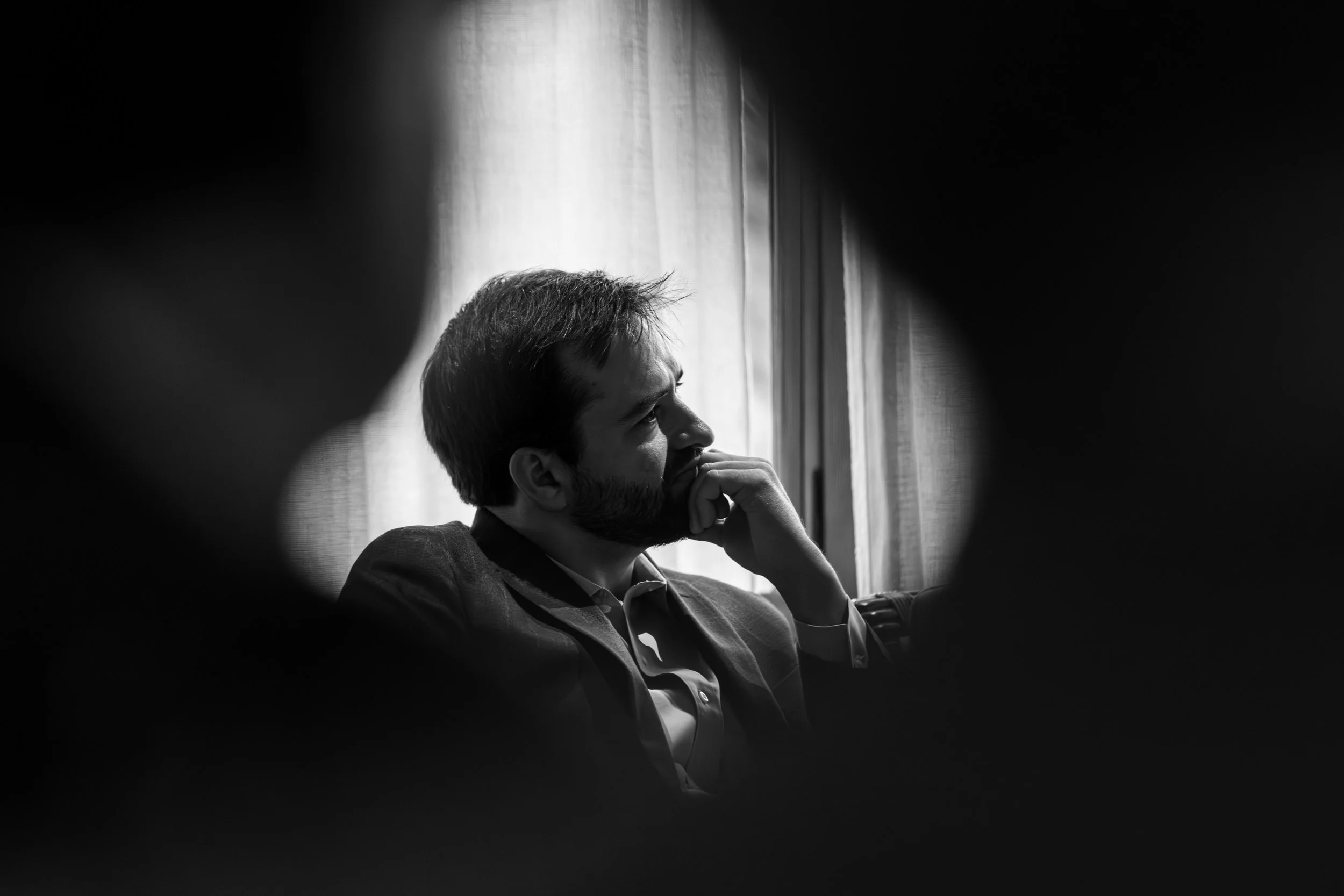 A black and white photo of a man sitting on a couch near a window, with his hand on his chin, viewed through a small circular opening.