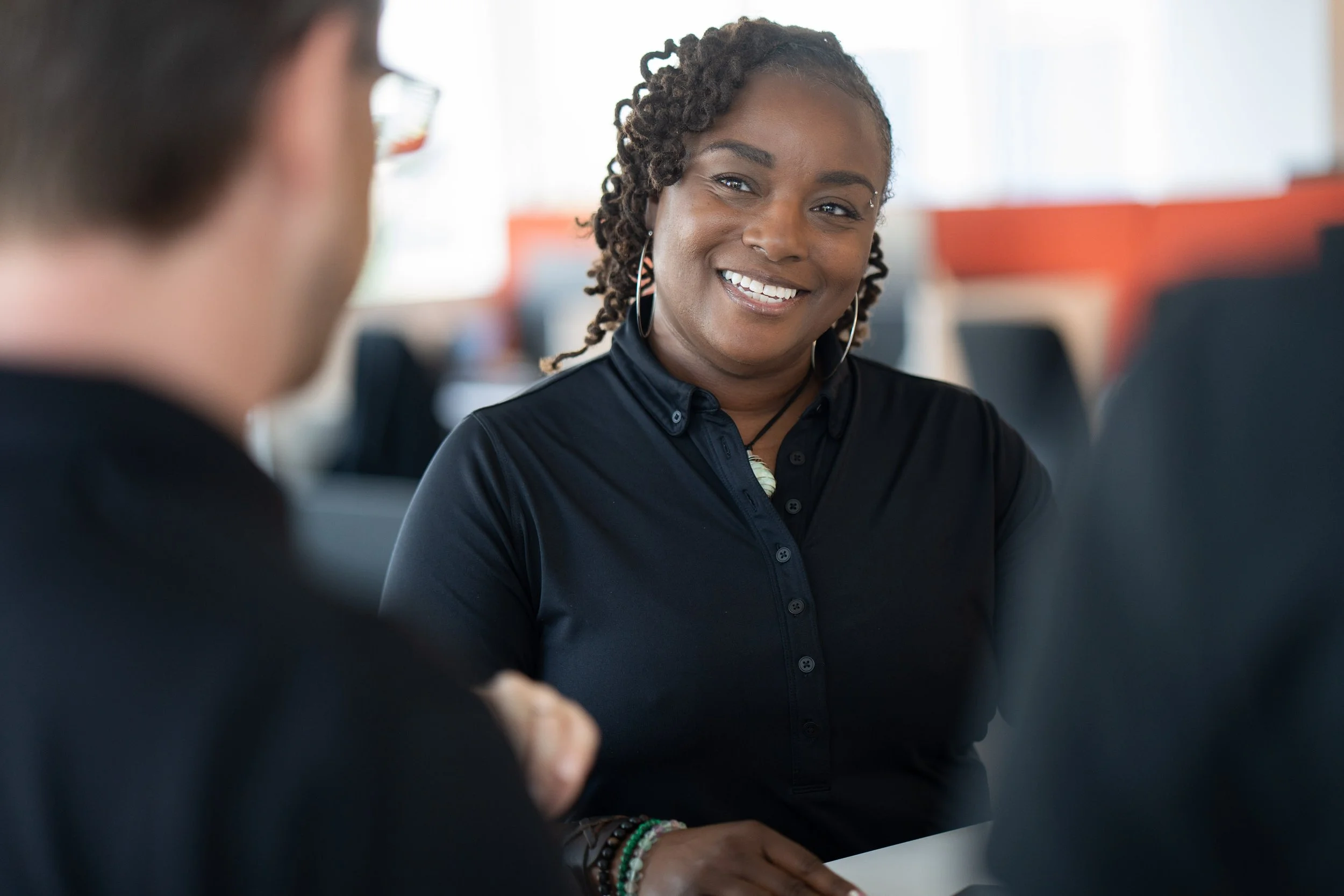 Smiling woman with earrings and jewelry, talking with a blurred person in a professional setting.