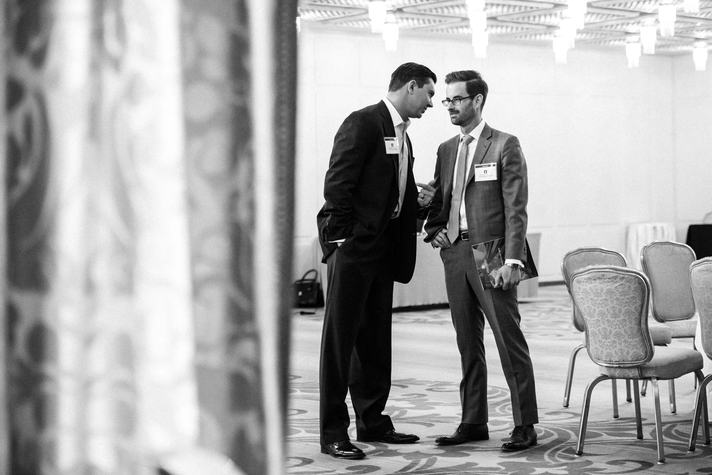 Two men in suits engaged in a conversation at a formal event or conference, with chairs arranged in a banquet style in the background.