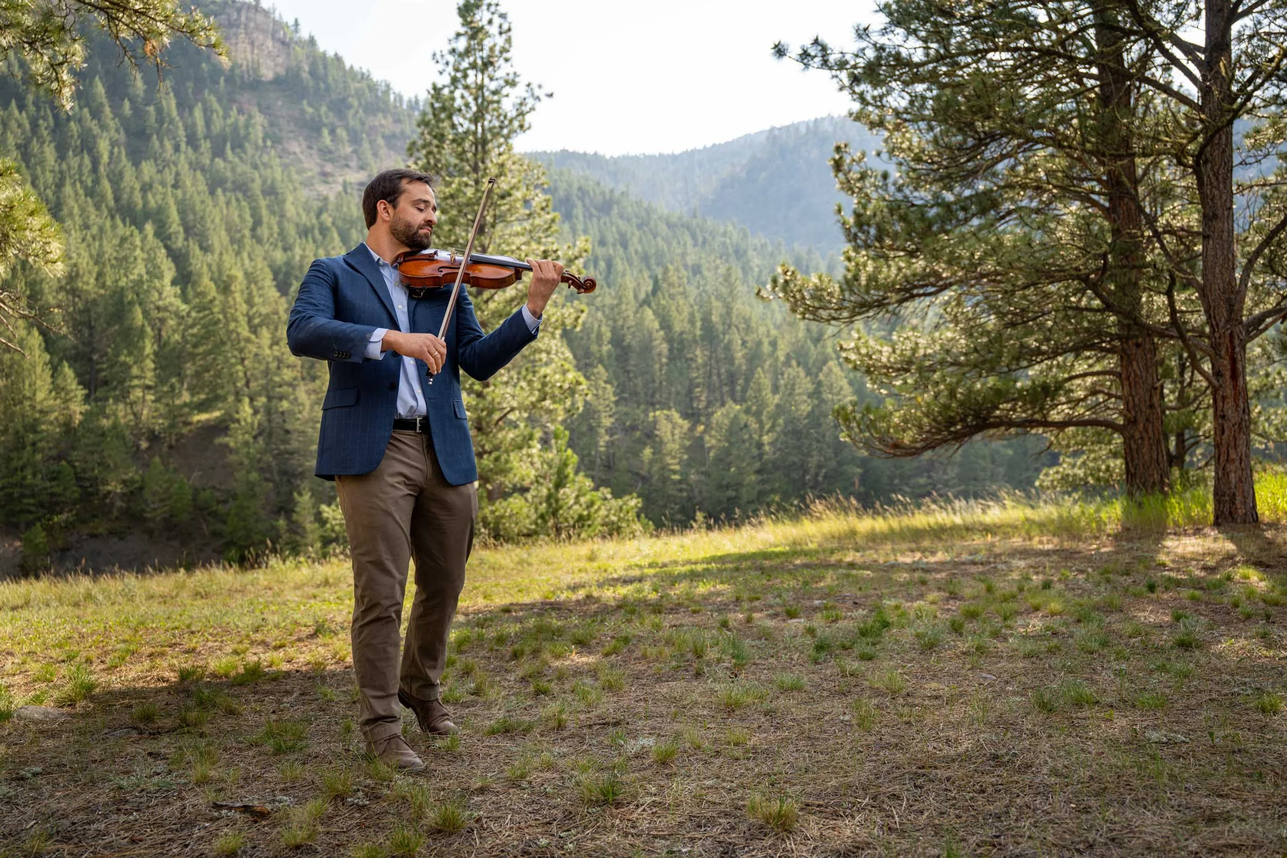 A man in a blue blazer and khaki pants playing the violin outdoors in a wooded mountain area during daytime.