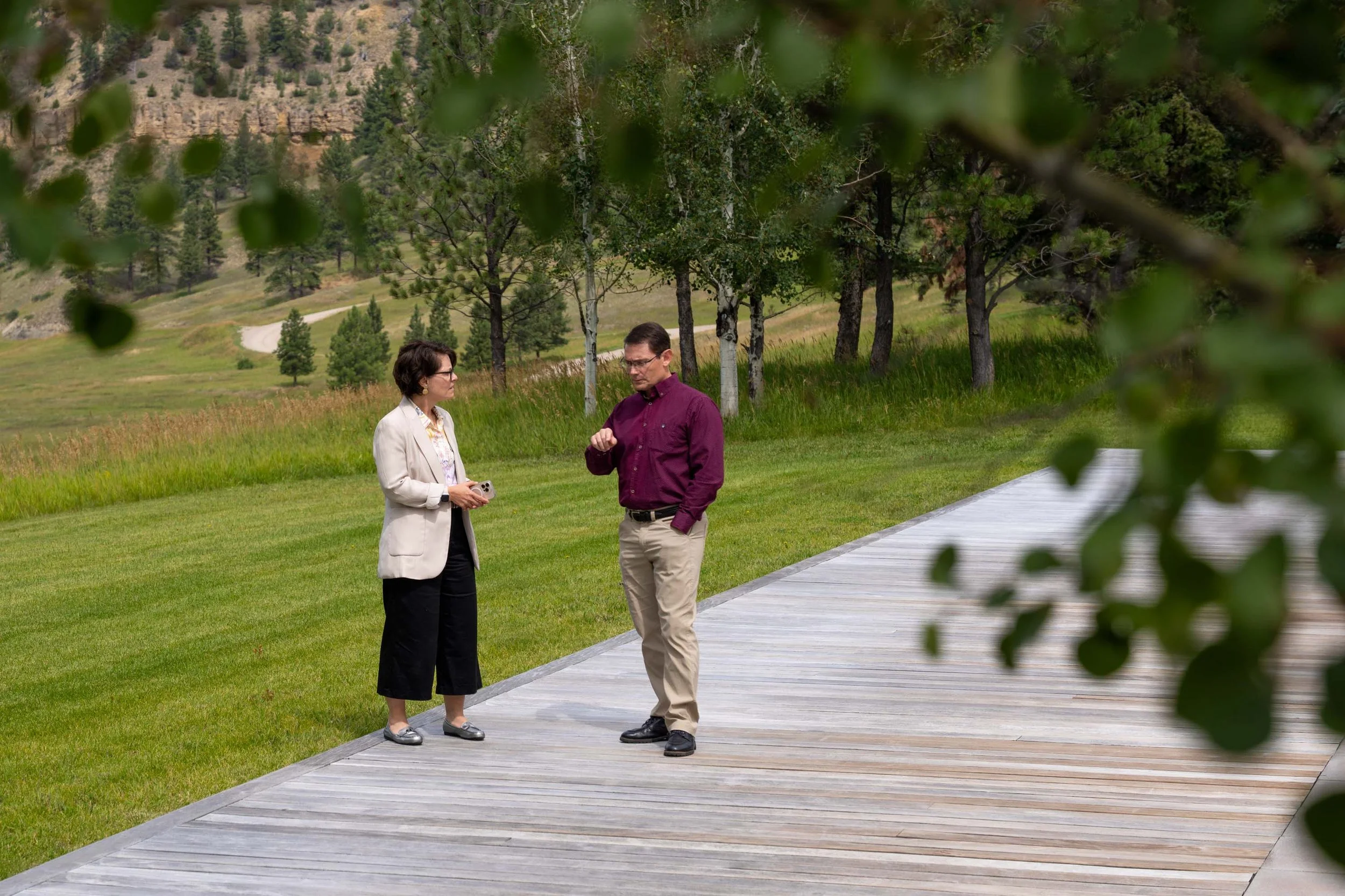 A man and a woman standing on a wooden pathway outdoors, engaged in conversation, with green grass, trees, and distant mountains in the background, partially framed by blurred leaves.