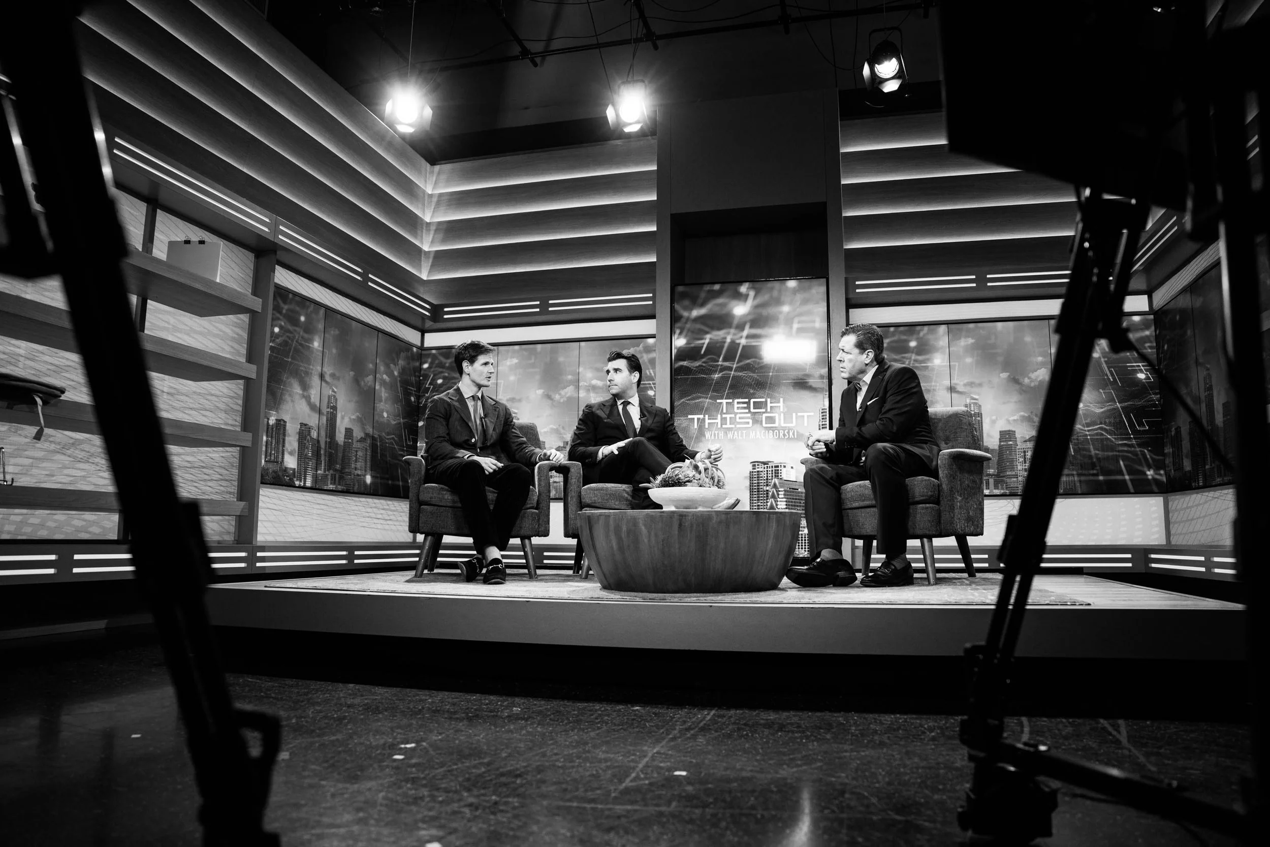A black and white image of a television studio set with three men seated on chairs engaged in conversation. The background features a cityscape with the words 'TECH THIS OUT' displayed on a screen. The photo is taken from beneath studio lights and ca
