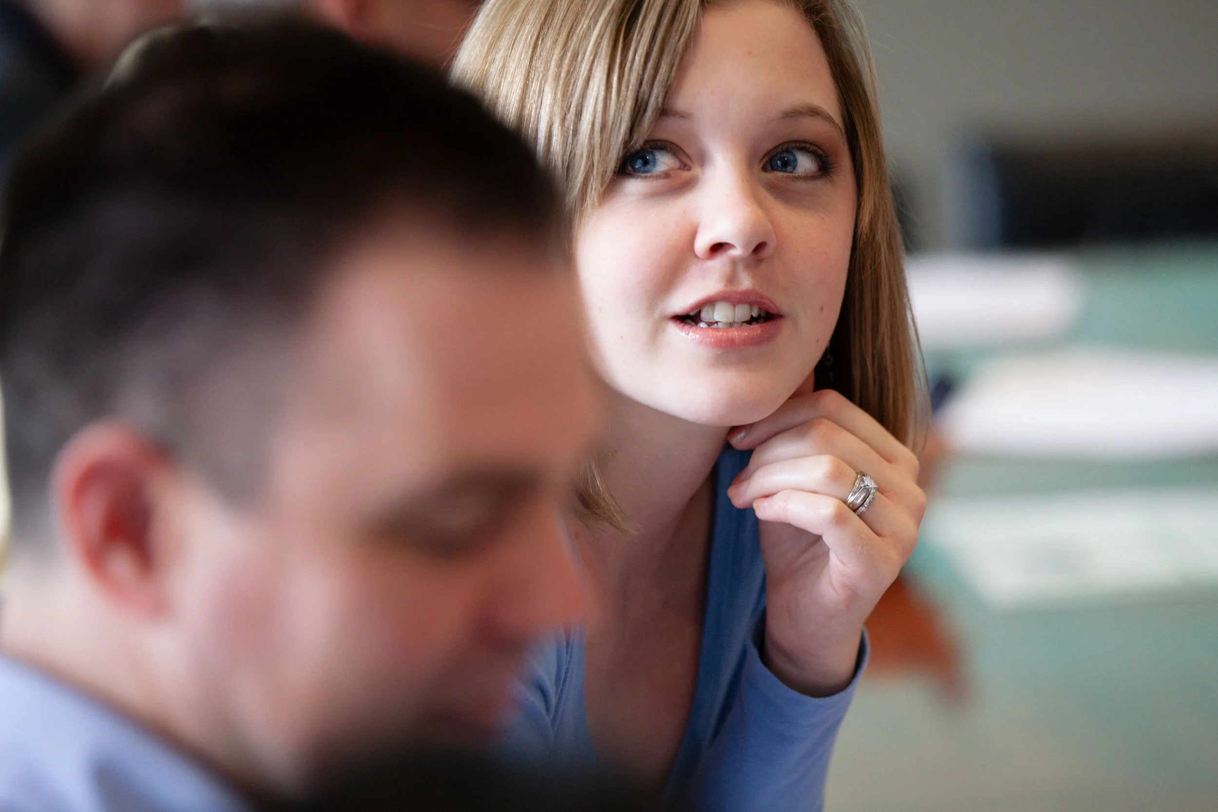 A woman with blonde hair and blue eyes, wearing a blue top, is engaging in a conversation, resting her chin on her hand, with a focused expression.