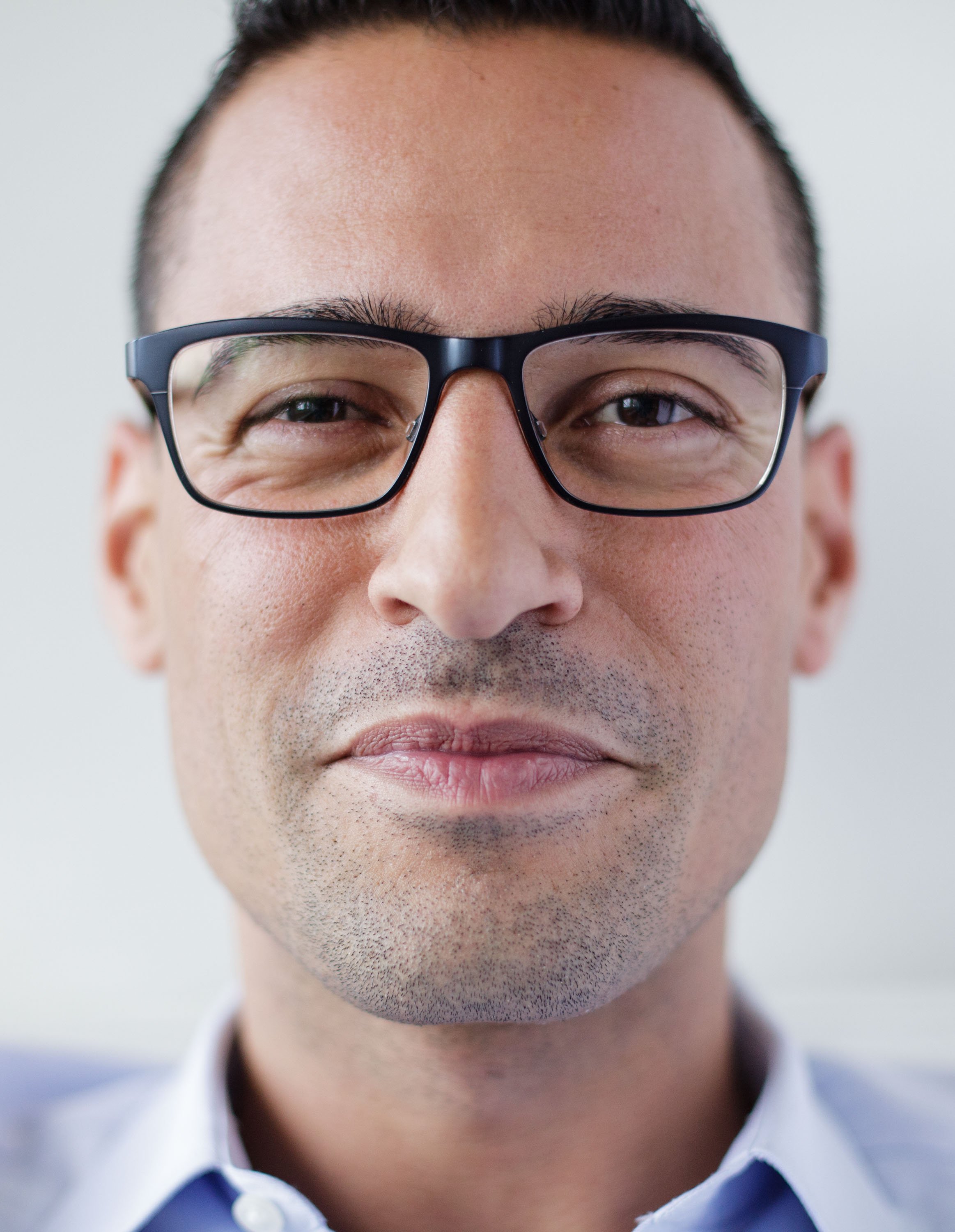 Close-up of a man wearing black-rimmed glasses with a slight smile, short dark hair, and wearing a white shirt with a blue collar, against a plain background.