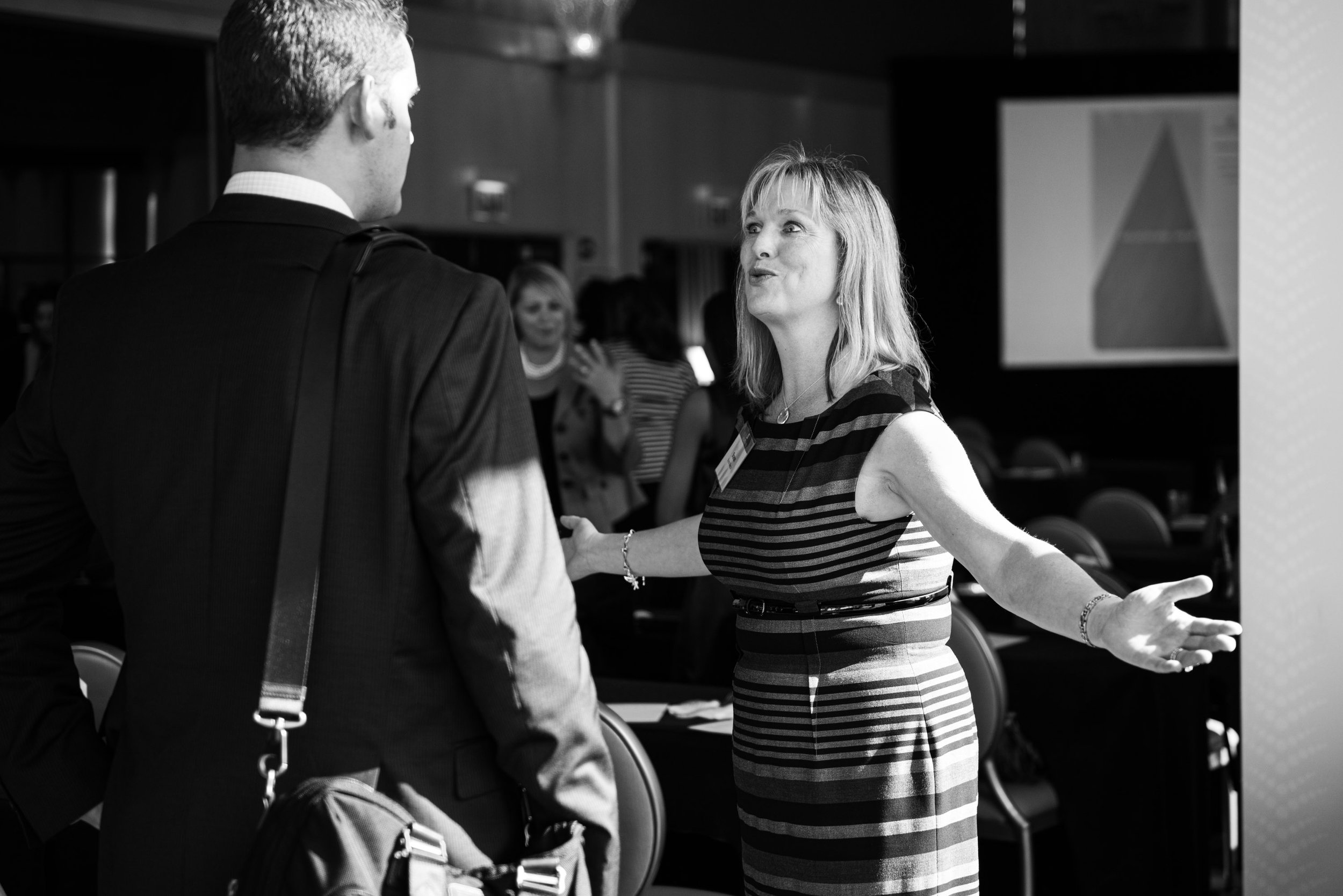 A woman with shoulder-length hair and a striped dress is engaging in conversation with a man wearing a suit, at what appears to be a professional event, with other people in the background.