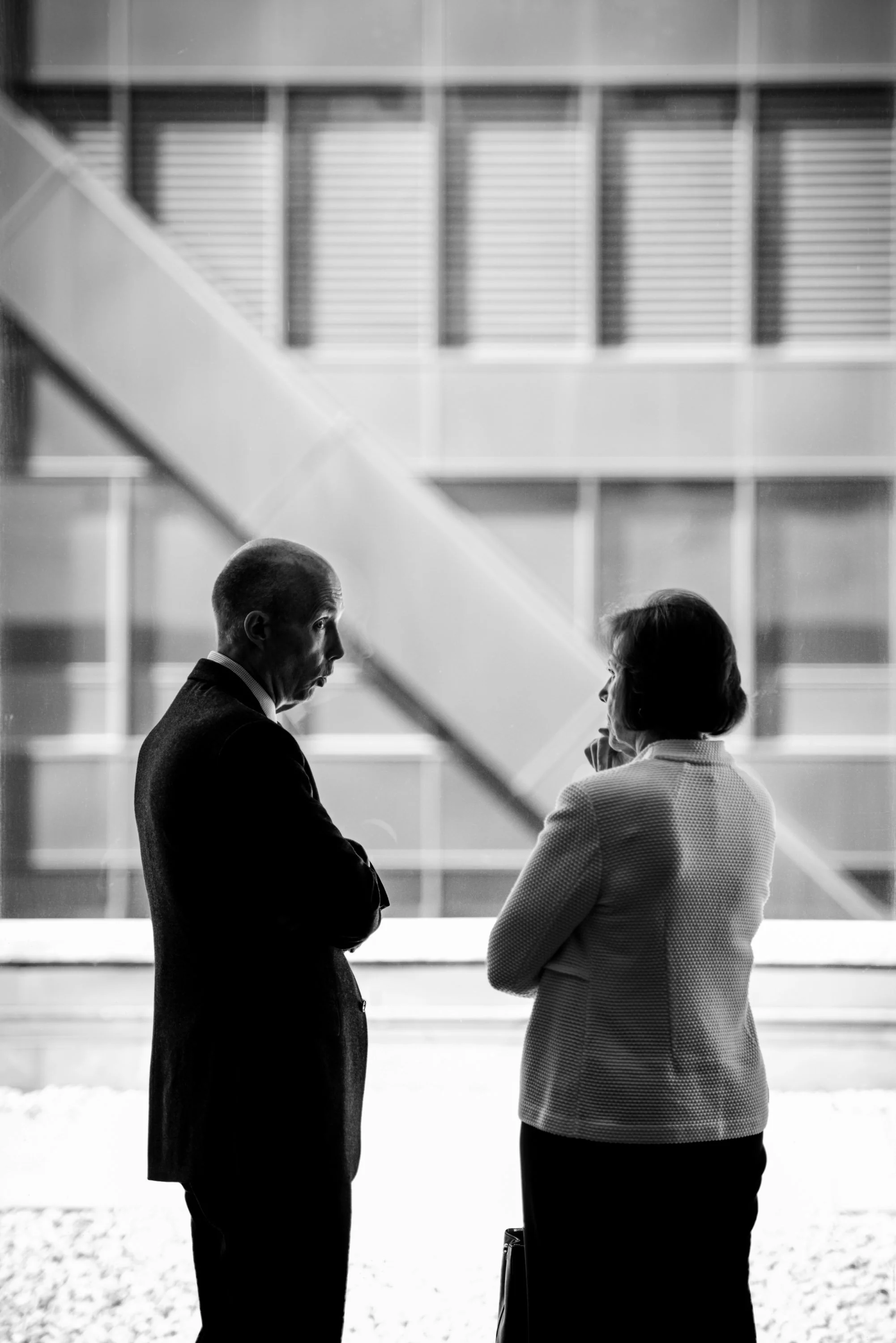 Black and white photo of two people in business attire standing and talking to each other in front of an office building with large windows.