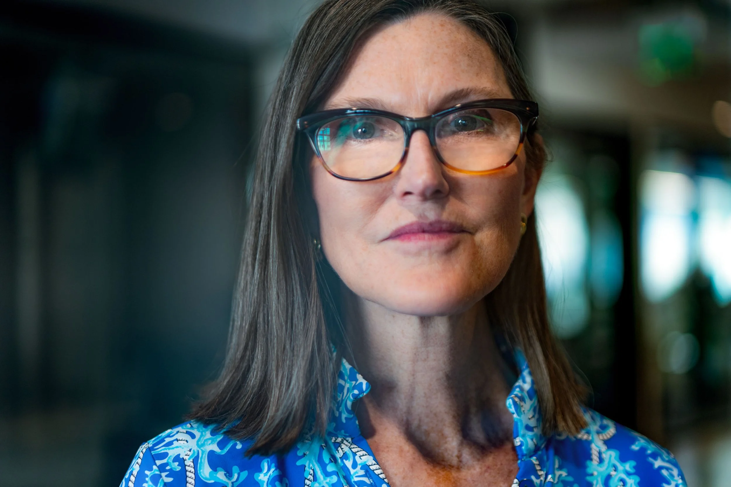 Close-up of a woman with long brown hair and glasses, wearing a blue patterned shirt.